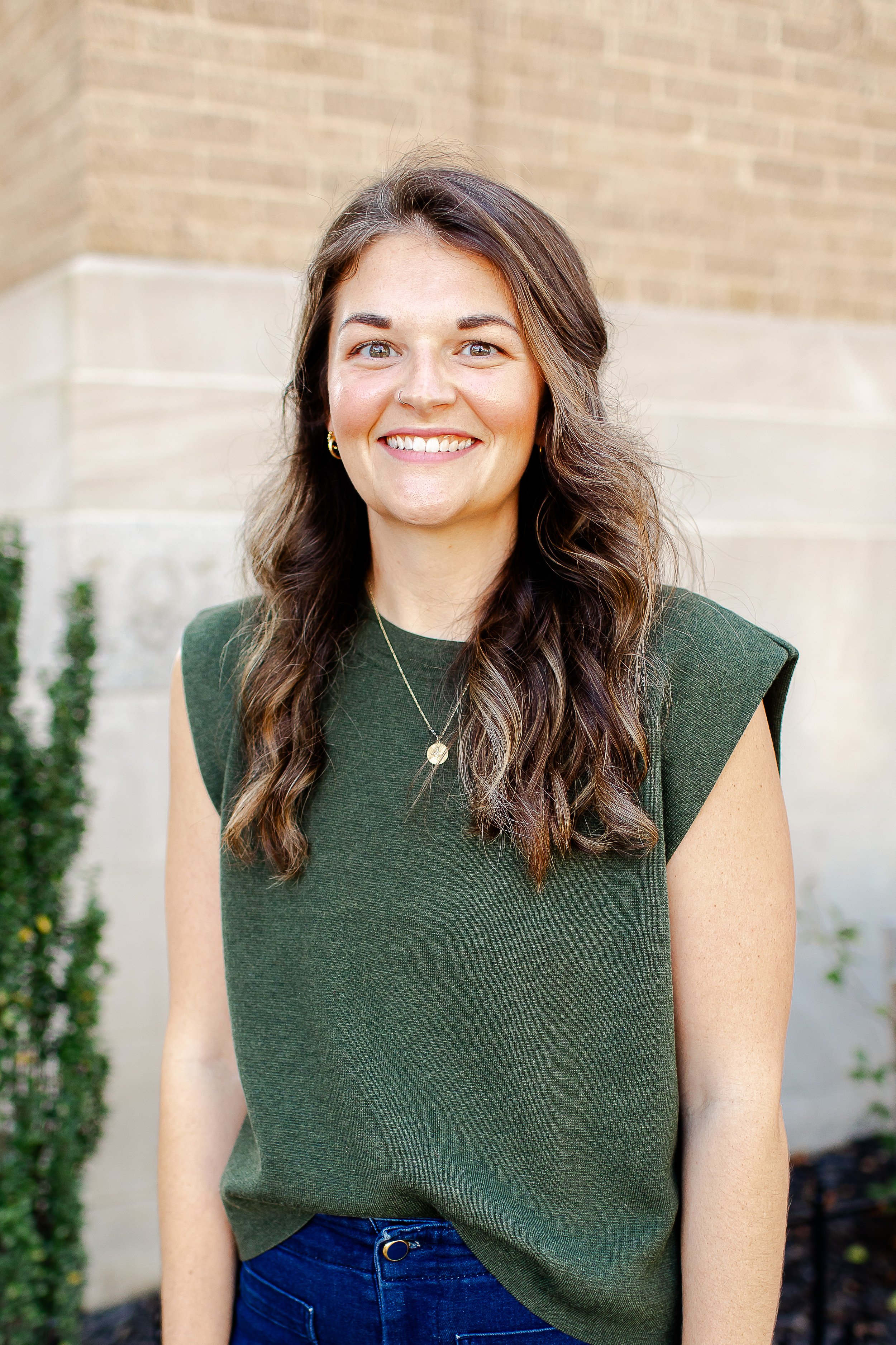 A smiling woman with long wavy brown hair and a nose ring, standing outdoors in front of a brick wall and plants, wearing a green sleeveless top and blue jeans.