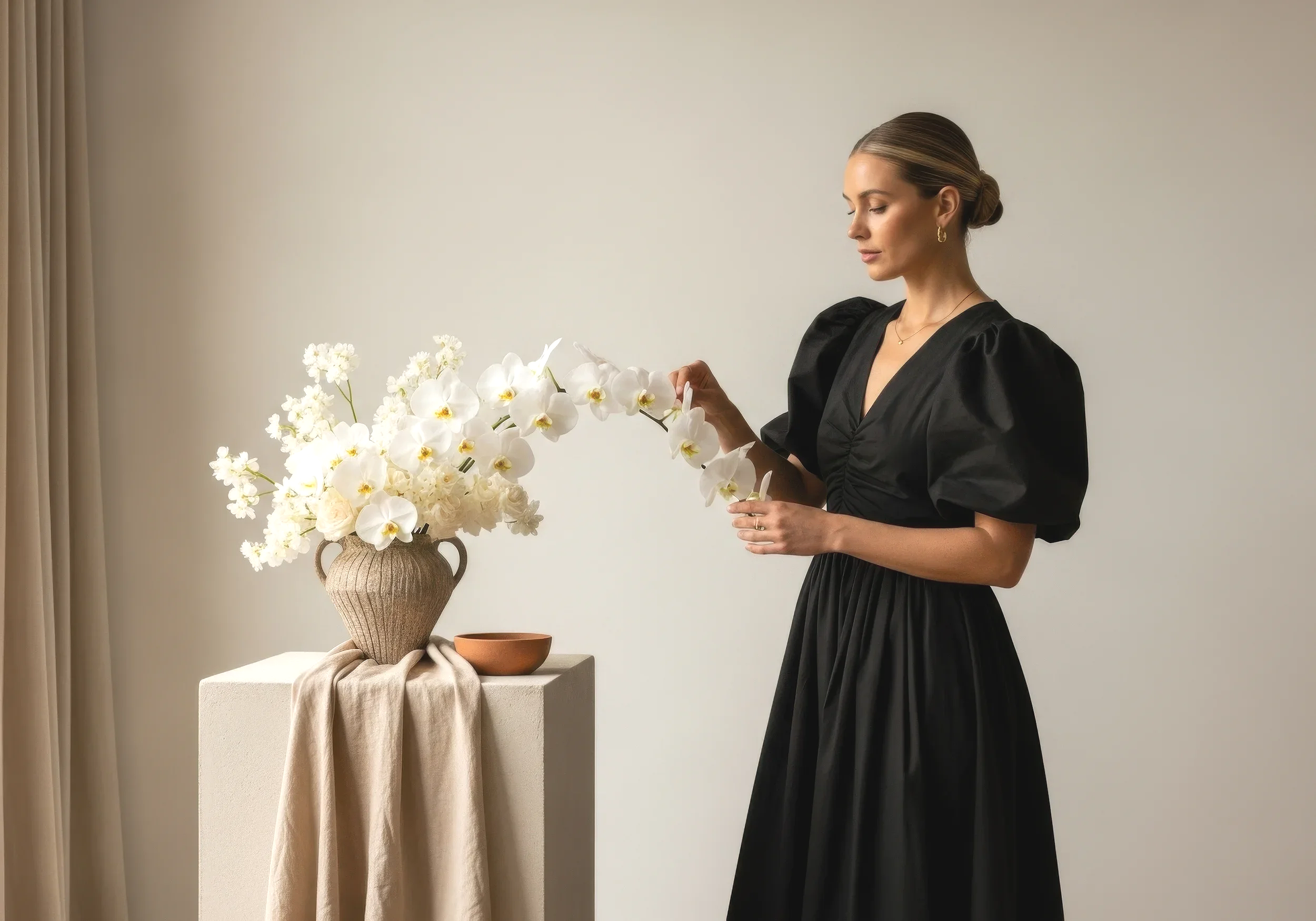 A woman in a black dress standing beside a table with a large floral arrangement of white orchids and other white flowers in a beige vase, with a beige curtain and plain wall in the background.