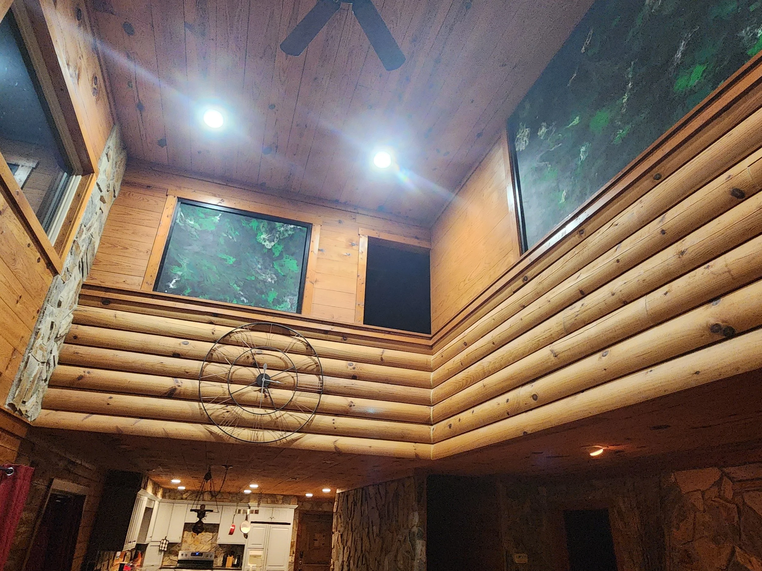 Interior view of a rustic cabin ceiling with wooden logs, including a ceiling fan, and windows showing greenery outside, with a kitchen area visible below. construction problem solve fill open space