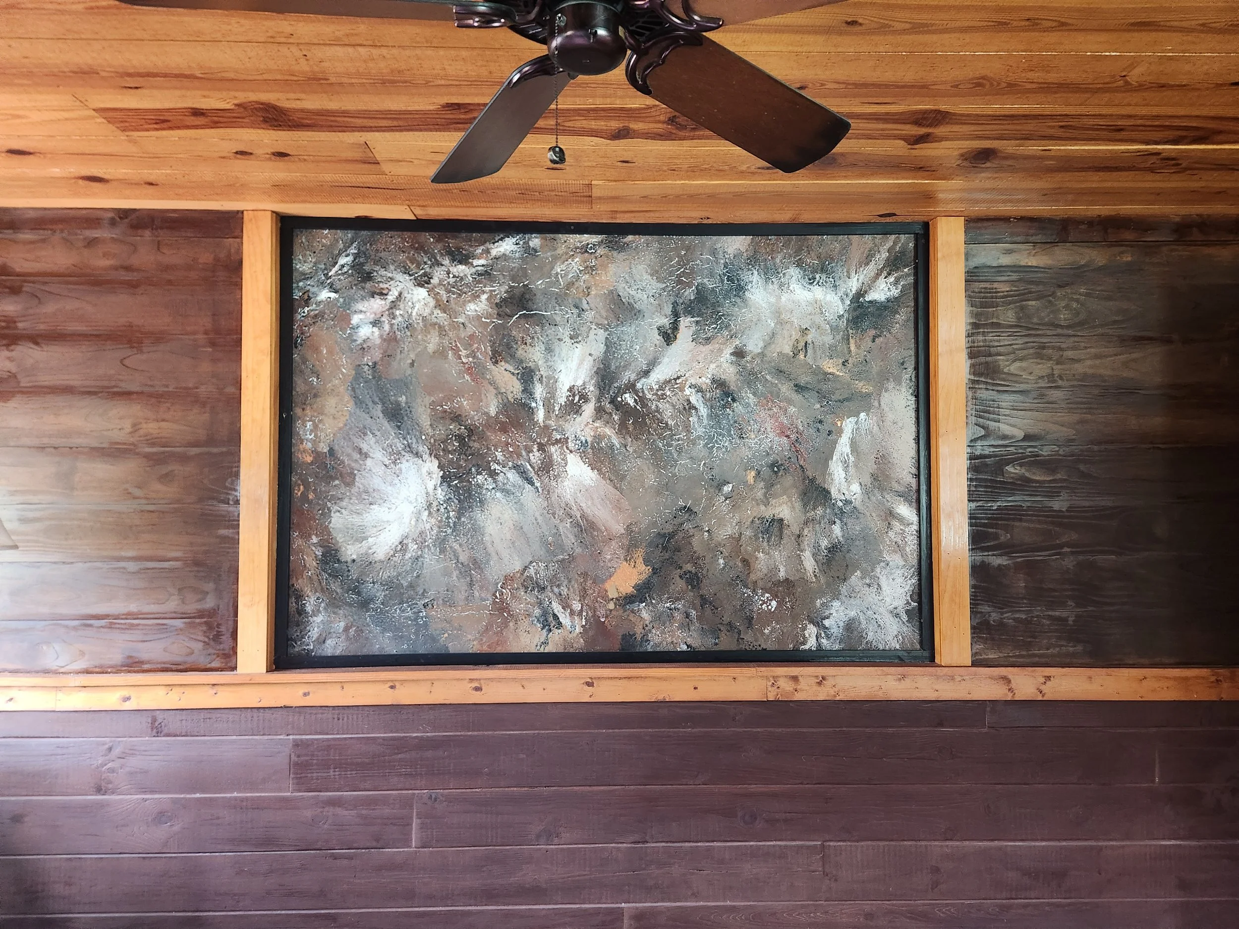 Photo of a portion of a room with wooden walls and floor, framing a painting or print of a natural landscape with trees, rocks, and a cloudy sky, viewed from below with a ceiling fan above.