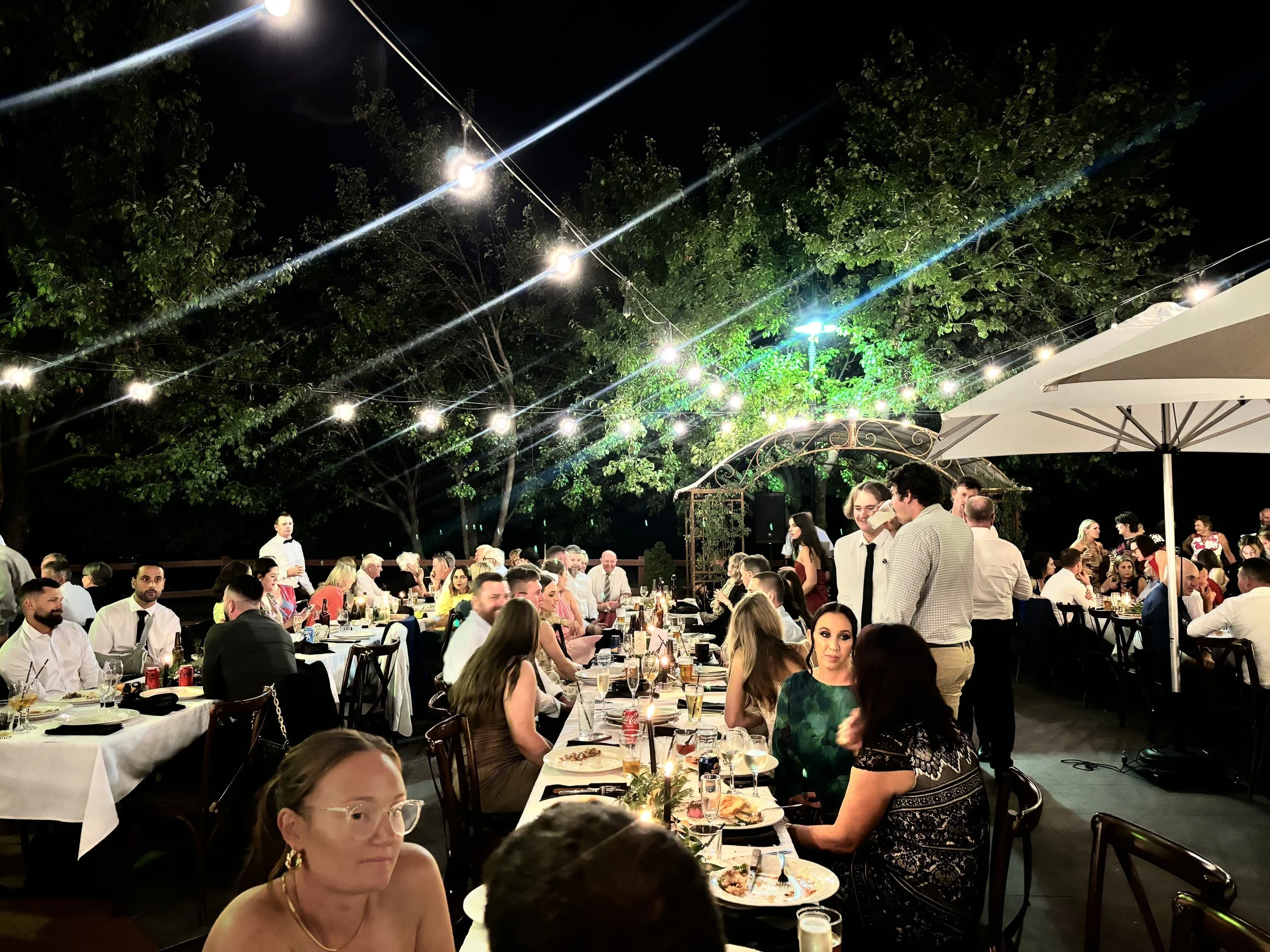 A bustling outdoor dining event at night with many people seated at tables under string lights and trees, celebrating around food and drinks.