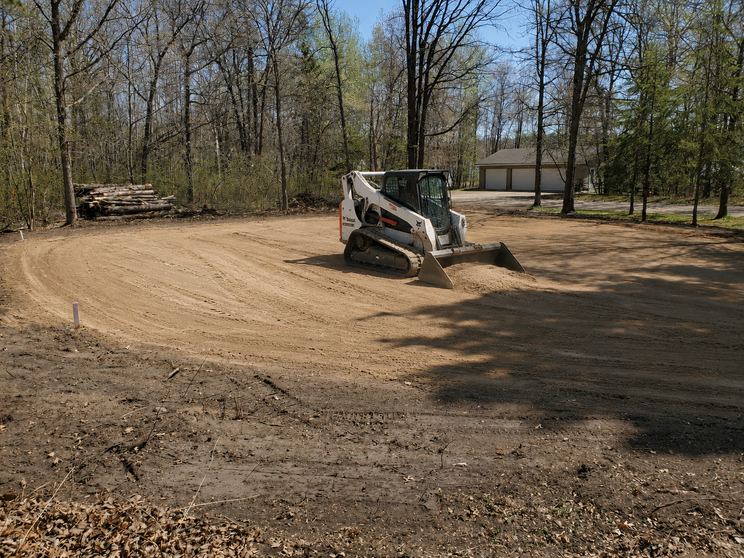 A small skid-steer loader working on a dirt construction site surrounded by trees and a residential garage in the background.
