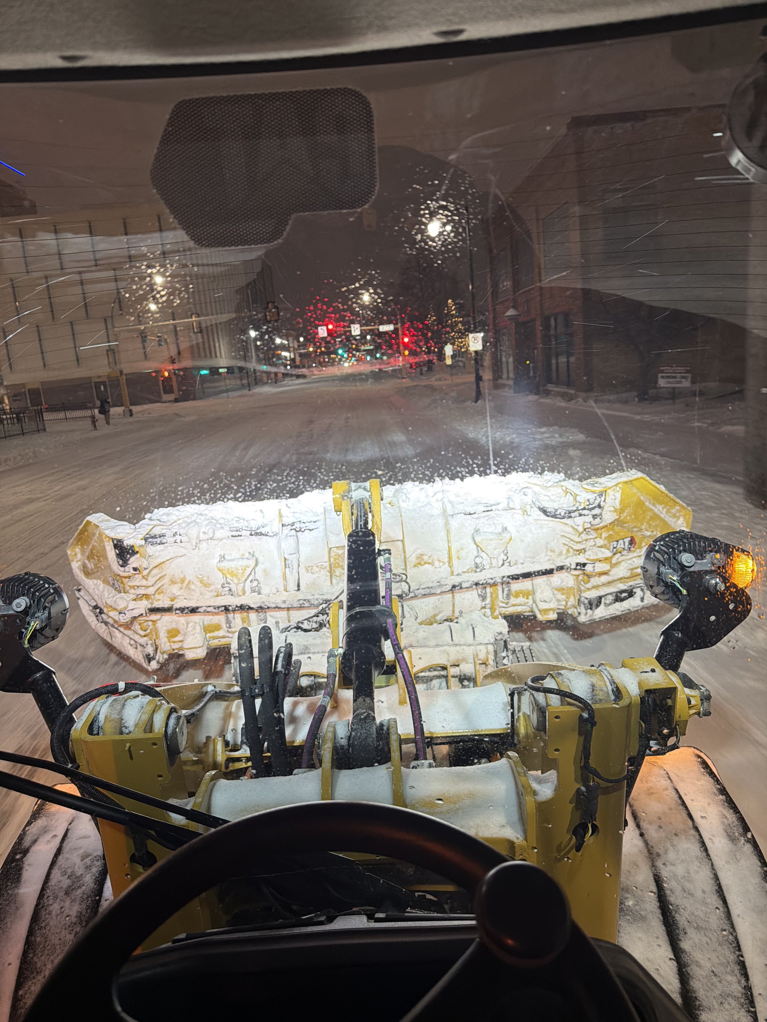 View from inside a snowplow vehicle at night, showing the snowplow blade clearing a snow-covered street with traffic lights and buildings in the background.