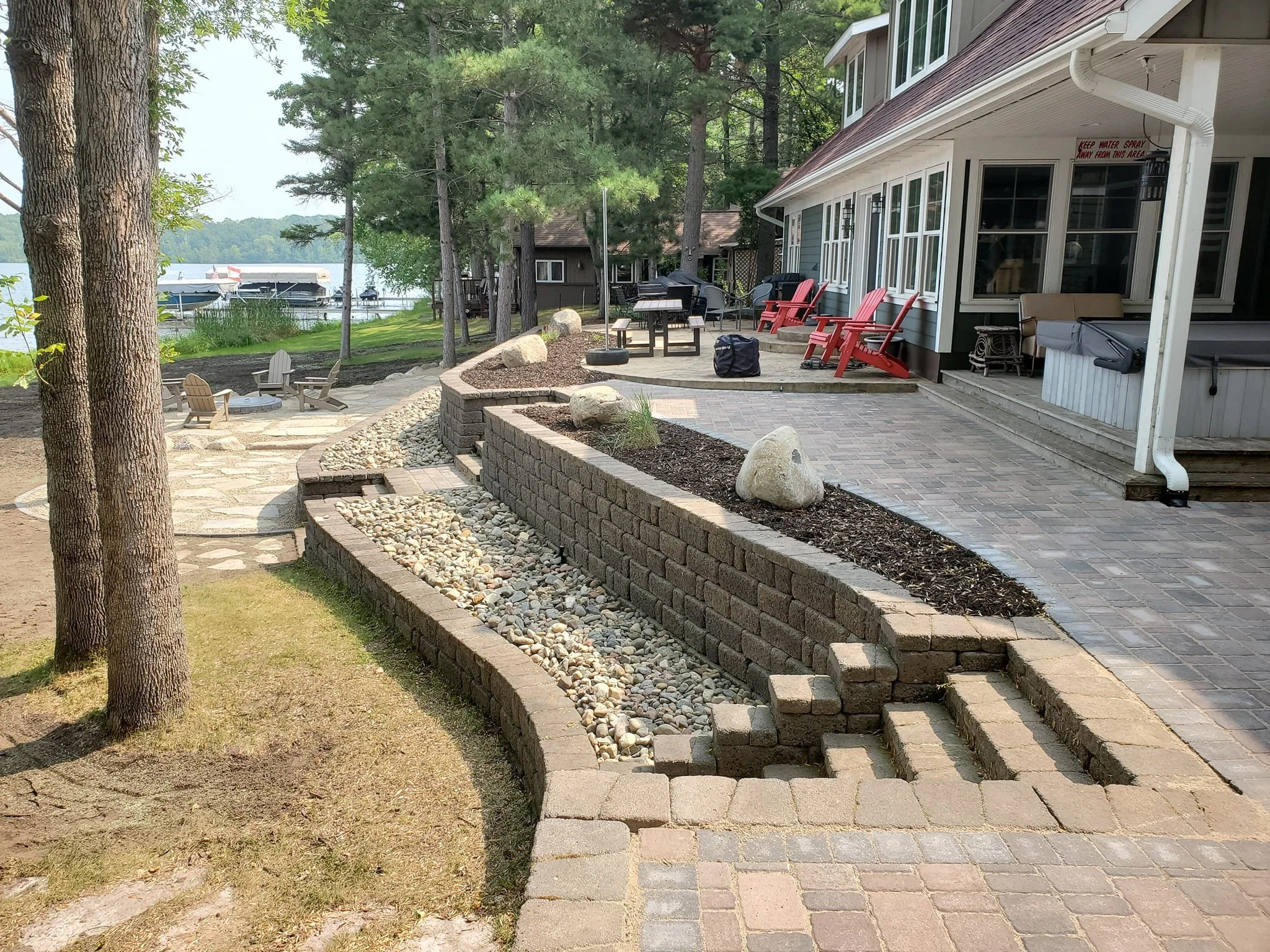 A backyard patio with brick steps and a retaining wall, surrounded by trees, with a view of the water and boats in the distance.