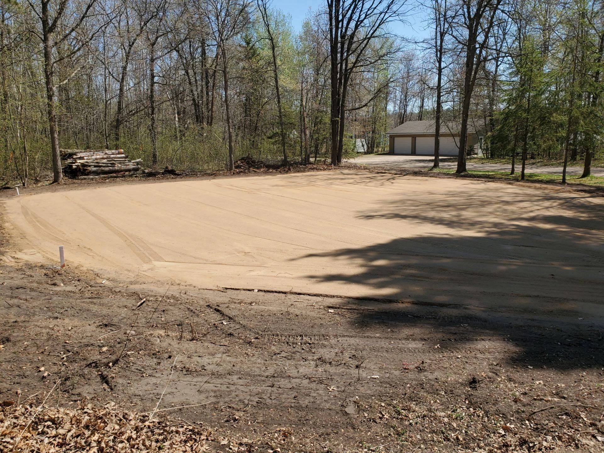 Empty construction site with a flat, cleared dirt area in foreground, surrounded by leafless trees and a detached garage in the background.