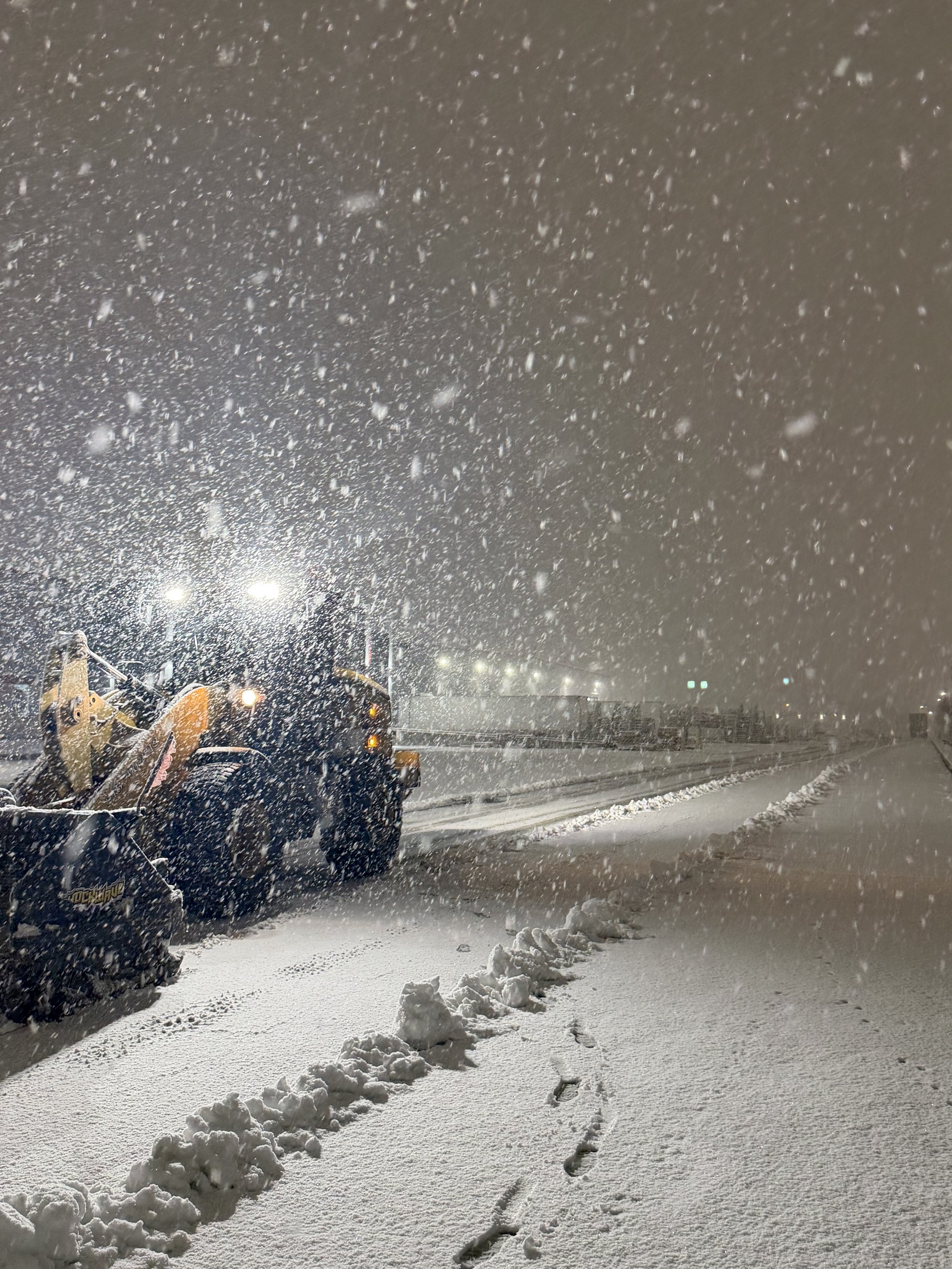 Snow-covered parking lot at night with a snowplow clearing the snow and footprints in the fresh snow.