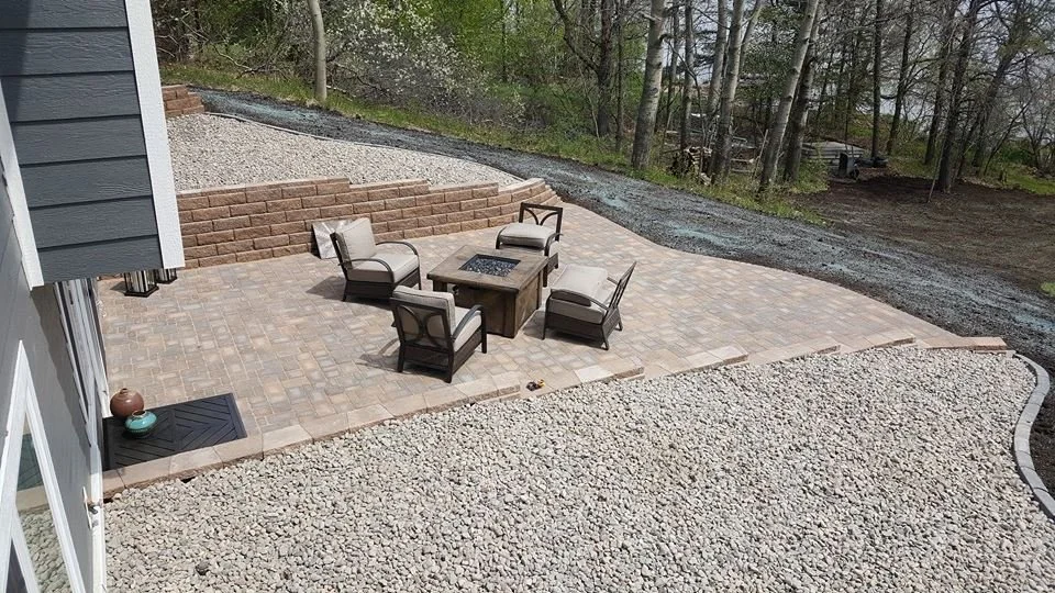 A backyard patio with four outdoor chairs around a fire pit table, surrounded by a gravel area and a retaining wall with steps leading up to a wooded area.