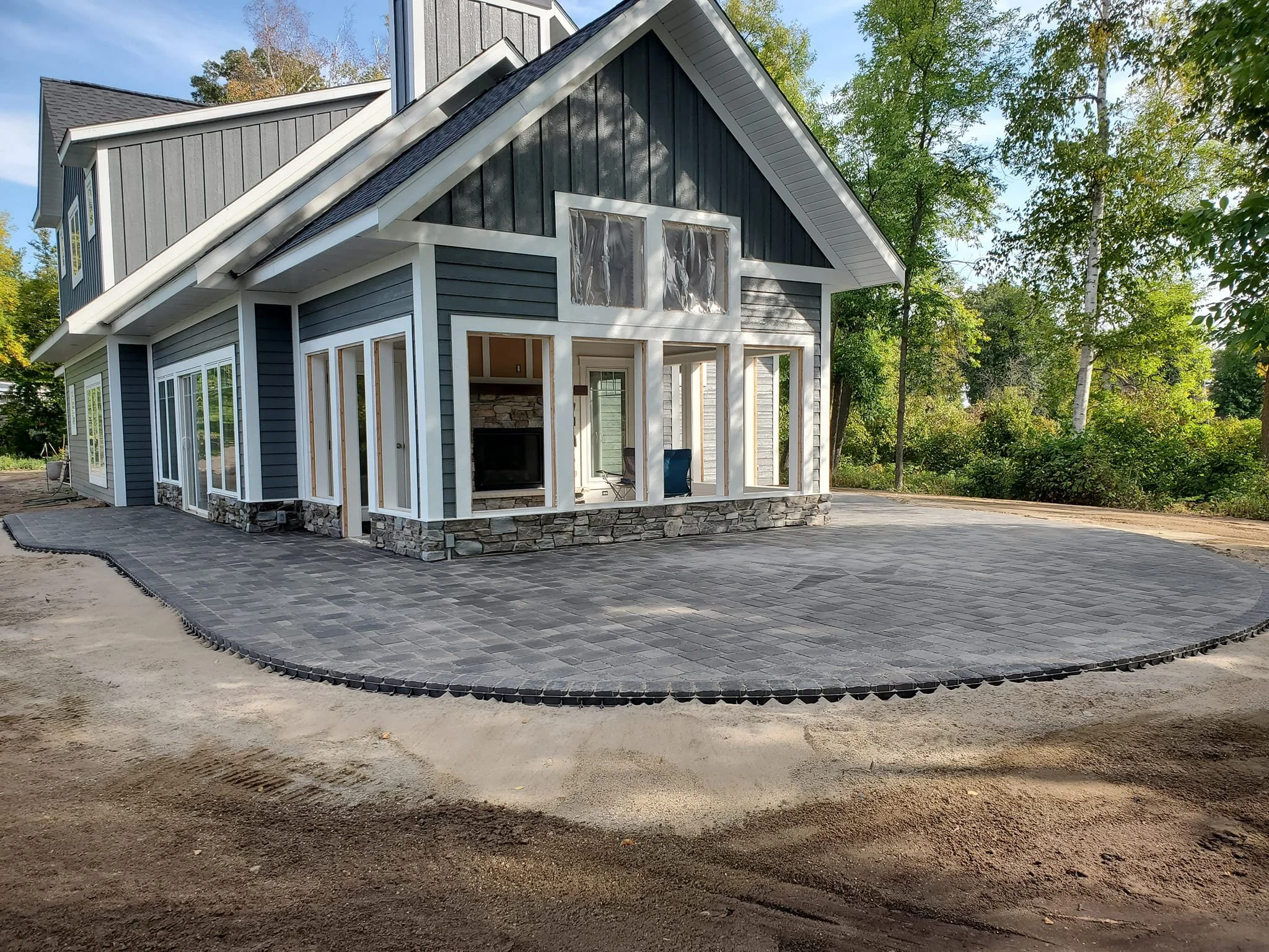 A house with gray siding and a stone foundation in the process of construction, with a paved area in front and surrounded by trees.