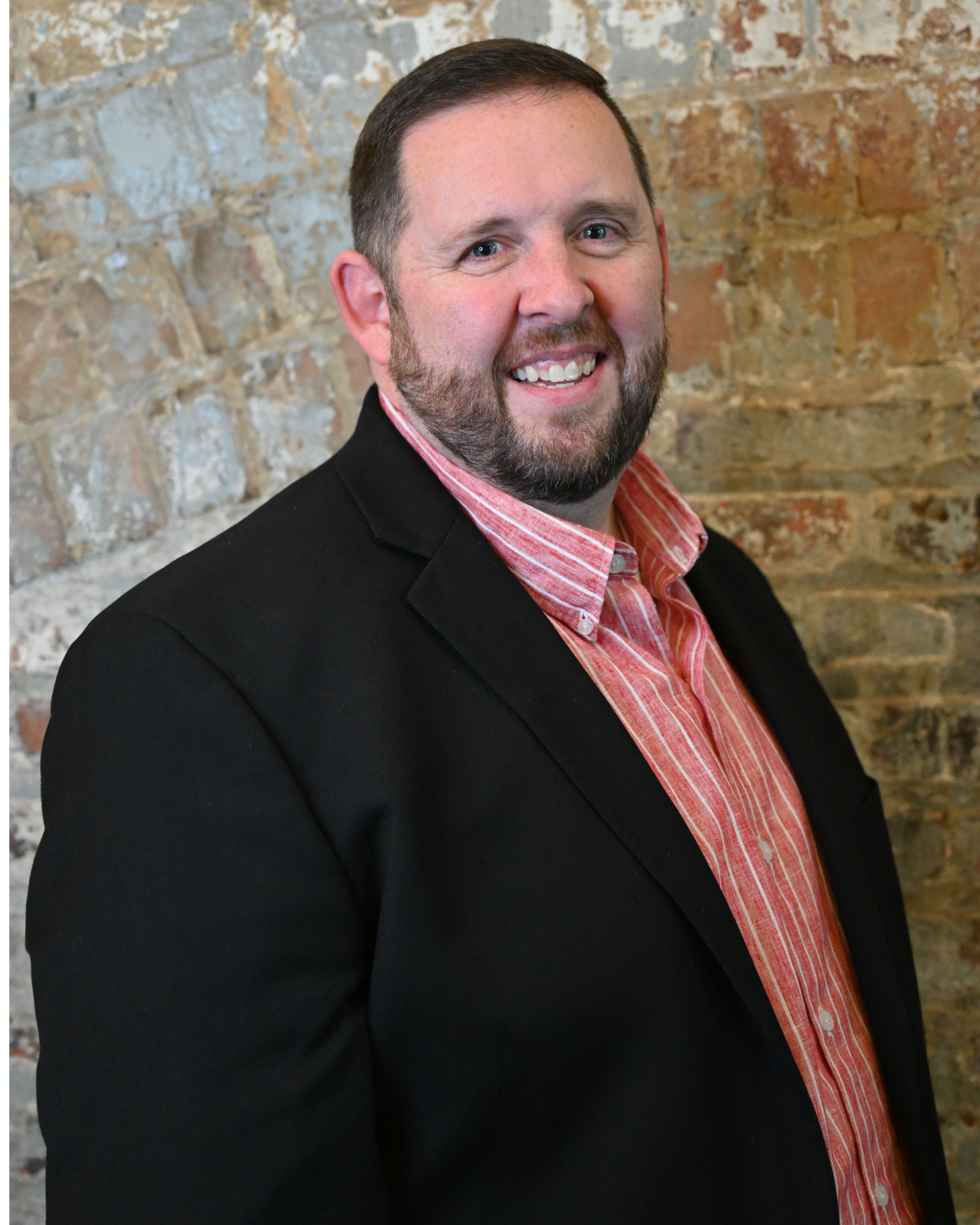 An image of Nathan with short dark hair and a beard wearing a black blazer and a red and white shirt, standing against a brick wall.