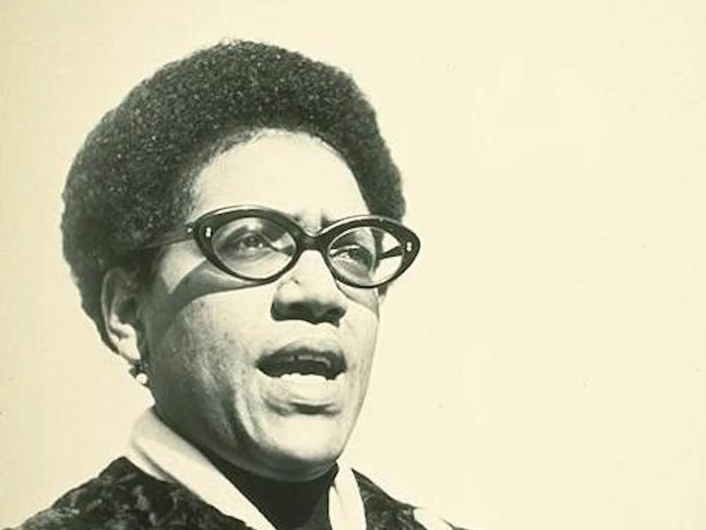 A black-and-white photograph of an African American woman wearing glasses, with an afro hairstyle, speaking or singing with her mouth open.