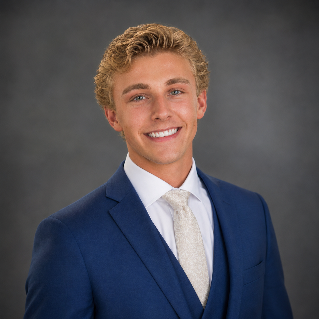 Portrait of a young man with blonde hair, blue eyes, and a big smile, wearing a navy suit, white shirt, and cream-colored tie, against a gray background.