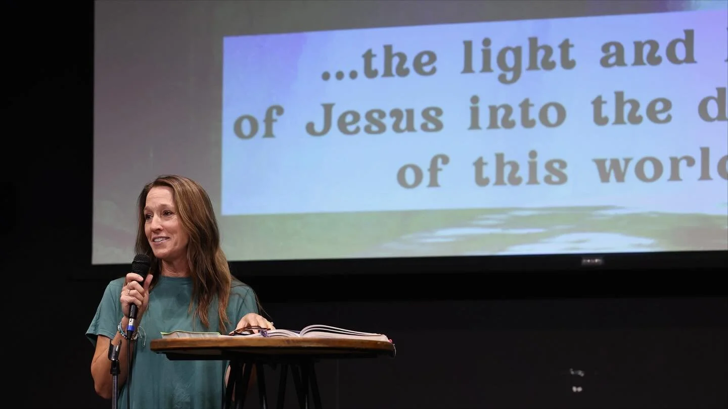 Rhonda Smith speaking into a microphone on stage, standing beside a small table with an open book. Behind her is a large screen displaying partially visible text about Jesus and light.