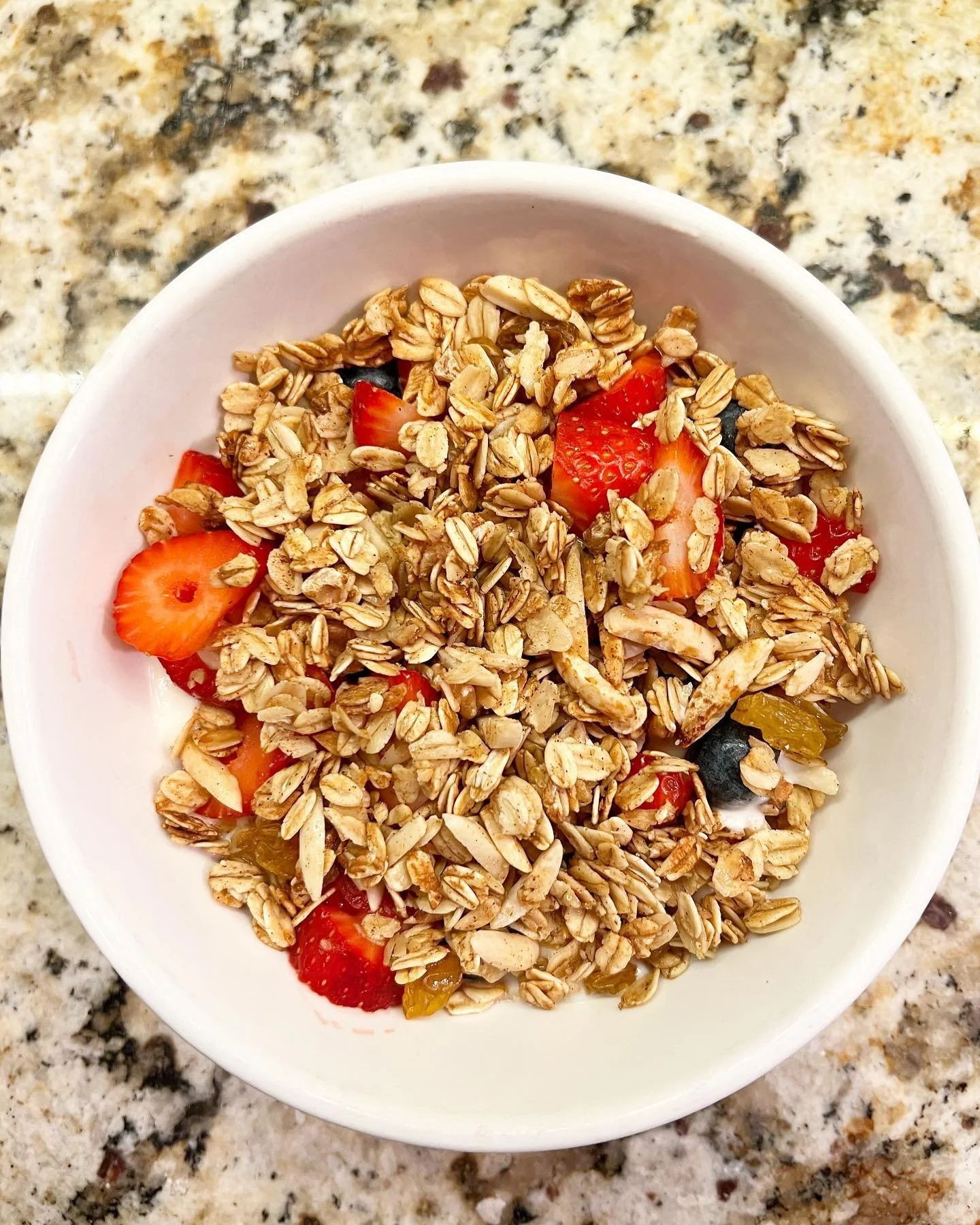 A white bowl containing cereal with oats, topped with sliced strawberries, blueberries, and pieces of yellow and red fruit on a granite countertop.
