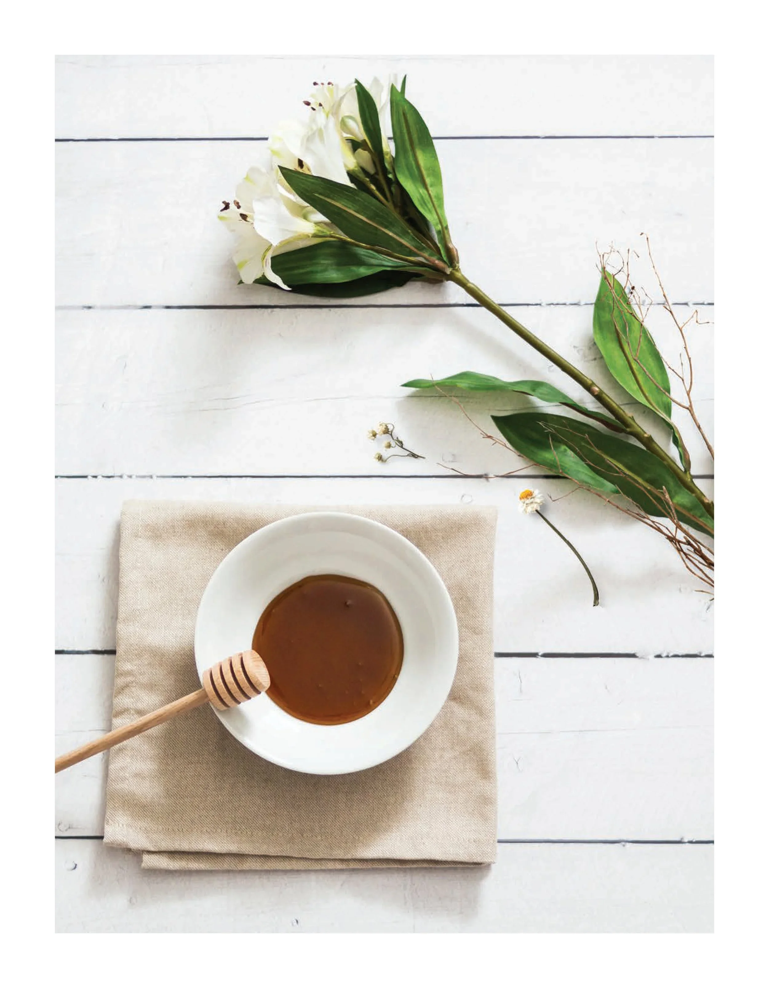 A white plate filled with caramel sauce and a honey dipper resting on a beige napkin on a white wooden table. Above the plate, there are a few sprigs of flowers and green leaves, and a branch with smaller leaves and flowers.
