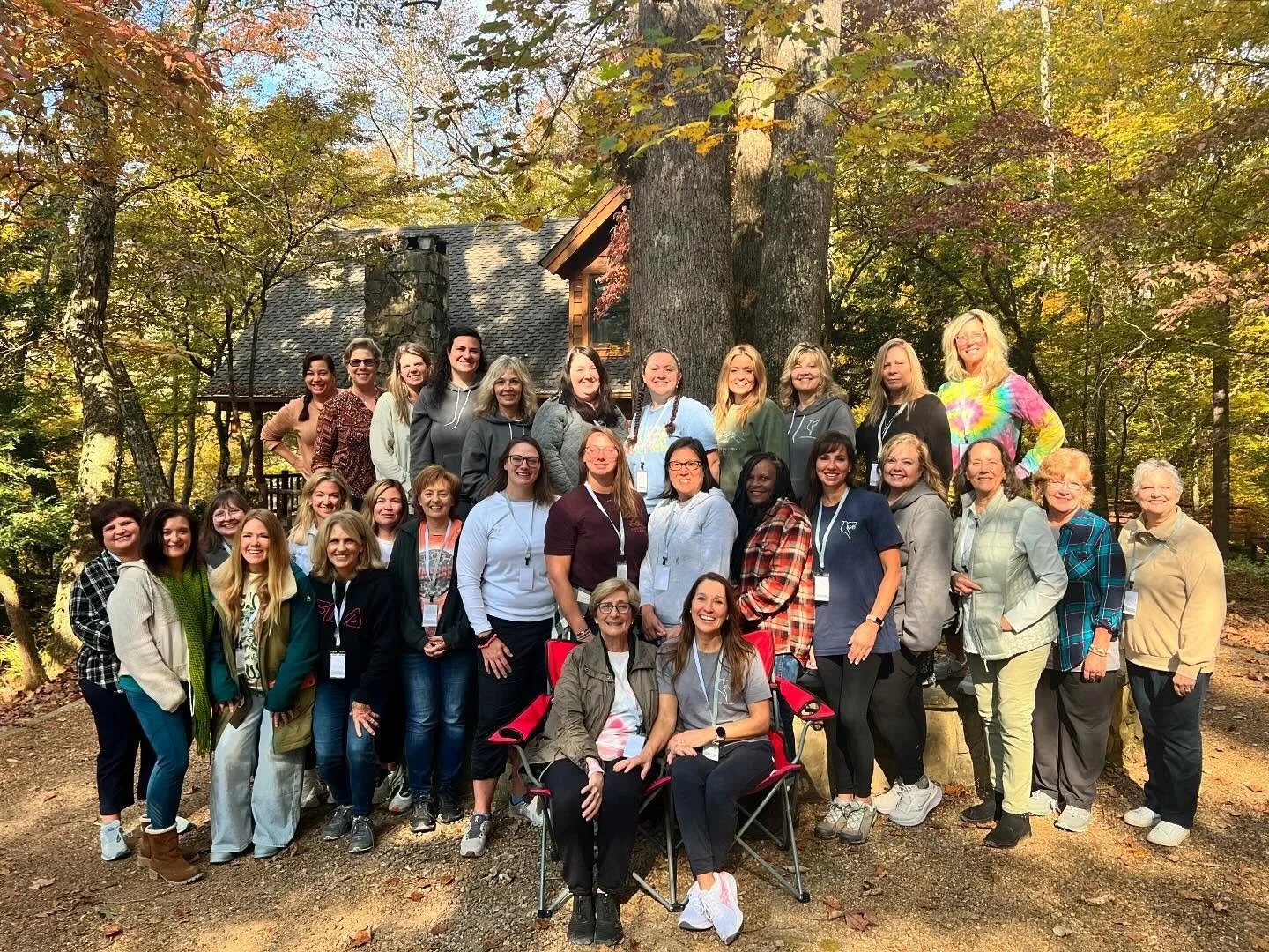 AWWG ladies at  first official Retreat gathered outdoors in a wooded area with autumn foliage, posing together for a photo in front of a large tree and a cozy cabin.