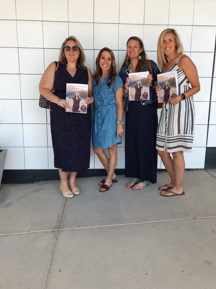 Four women standing against a white tiled wall, smiling and holding up copies of a book titled "Walk with God."