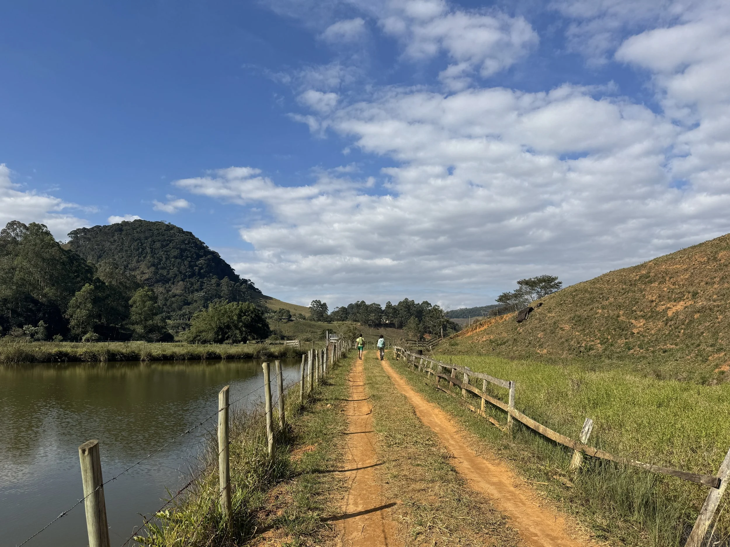 A dirt path running alongside a body of water, with a wooden and wire fence on either side, green hills and trees in the background, and two people walking along the path under a blue sky with scattered clouds.