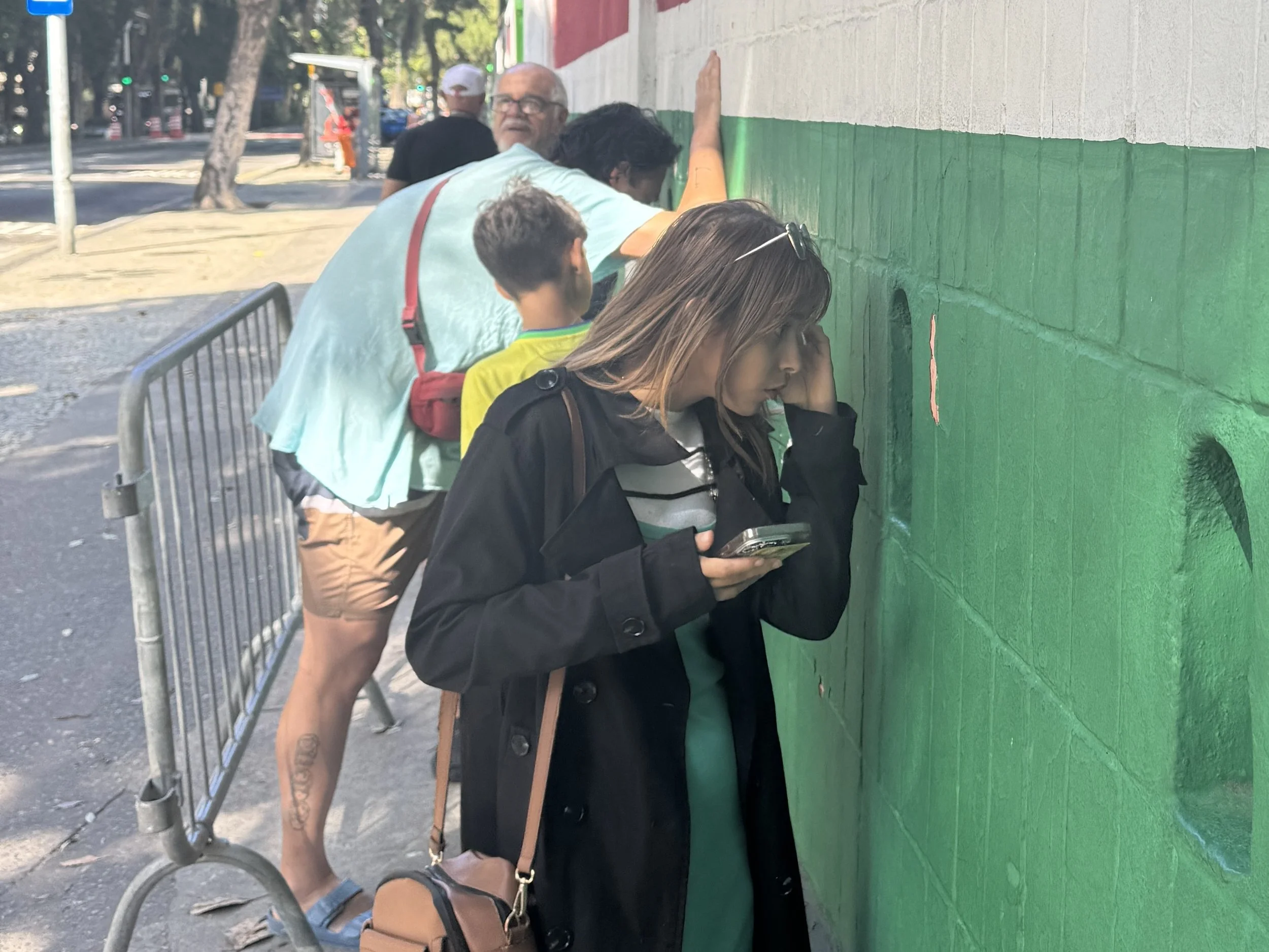 People creating a mural on a brick wall, with some individuals painting and others observing, behind a metal barricade.
