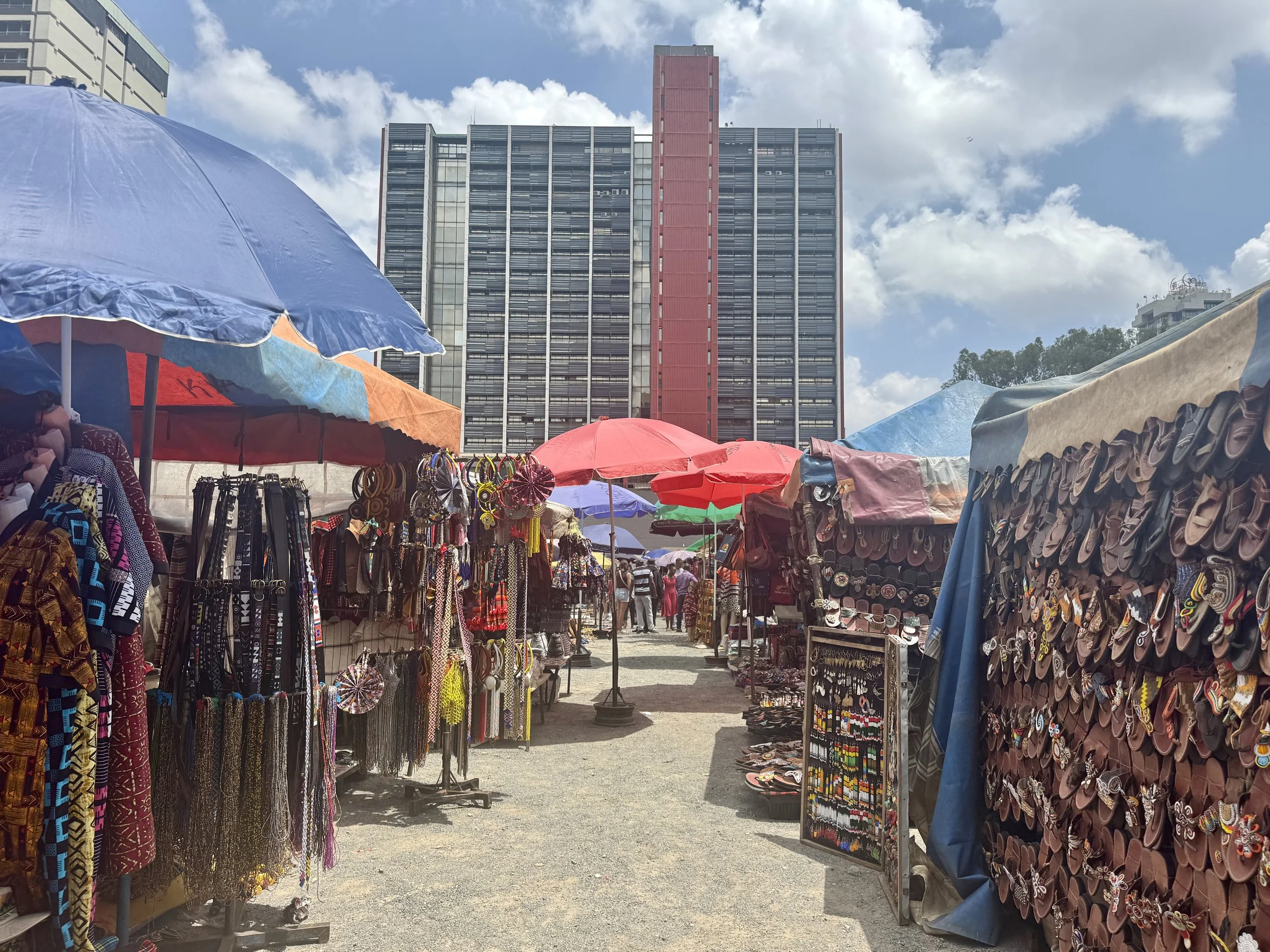 Market stalls selling accessories like belts, jewelry, and textiles, with colorful umbrellas and high-rise buildings in the background under a partly cloudy sky.