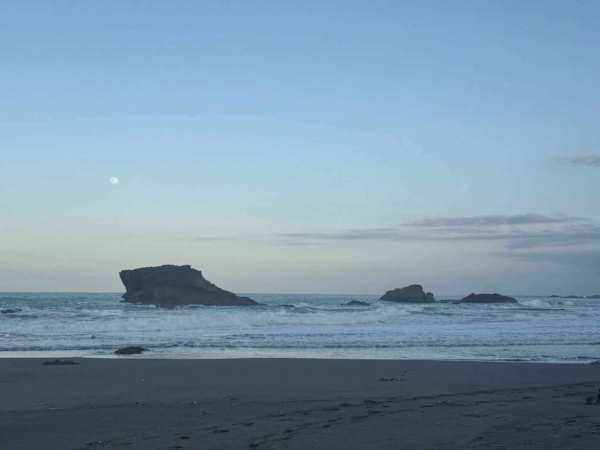 A beach scene during evening or early morning with rocks in the water, gentle waves, and the moon in the light blue sky.