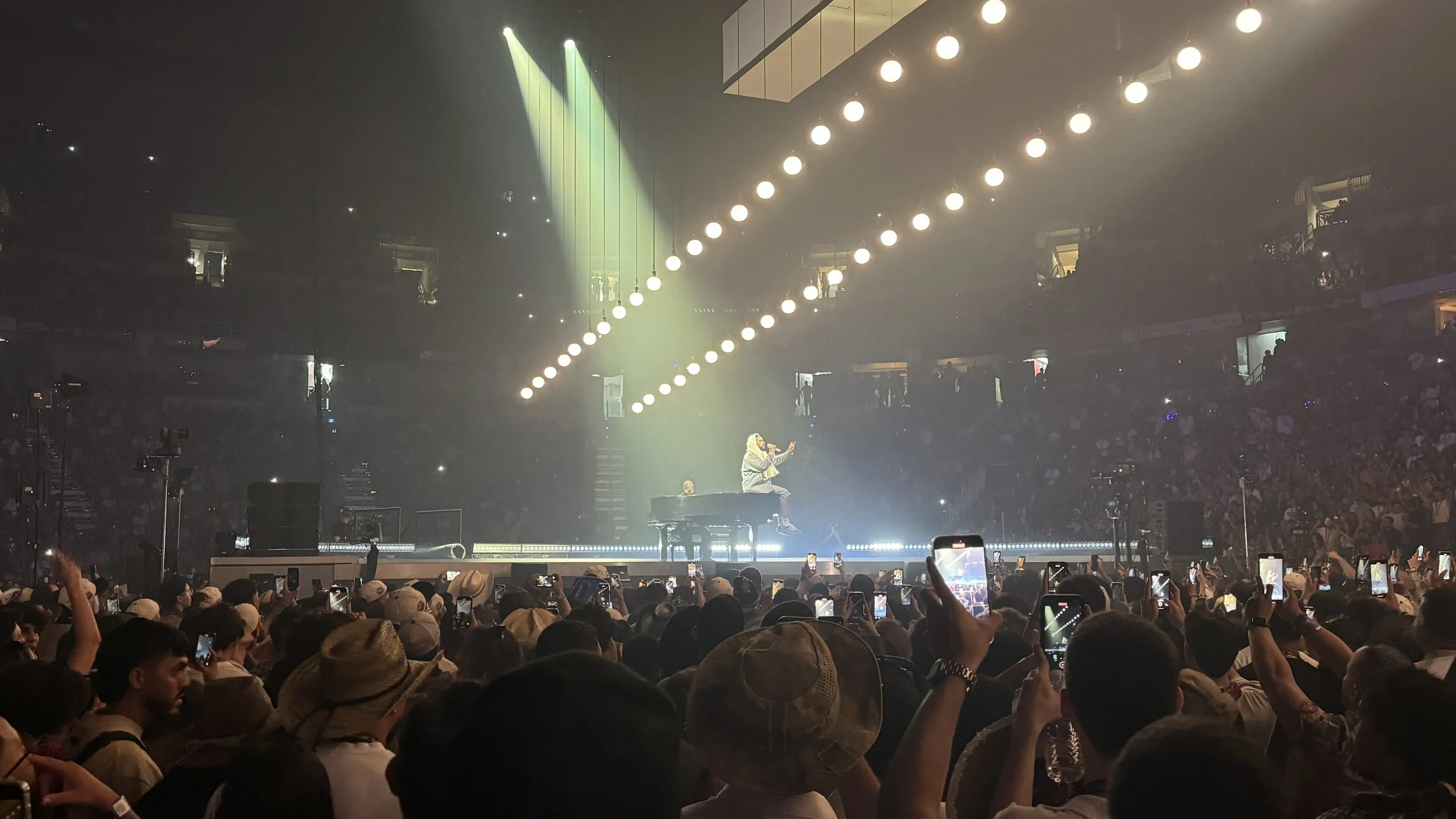 A concert scene with a singer sitting on a piano on stage, surrounded by a large crowd of fans. The stage is lit with bright lights, and many audience members are capturing the moment with their phones.