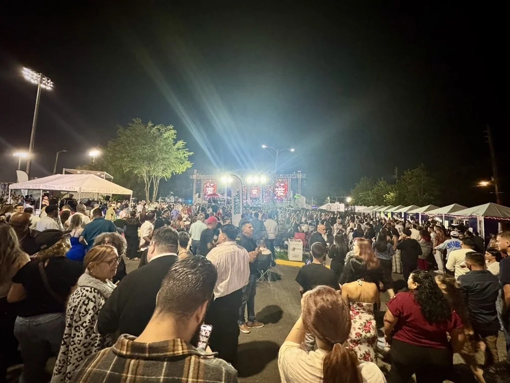 A crowded outdoor concert at night with many people standing and facing a brightly lit stage with performers and video screens, surrounded by tents and trees.