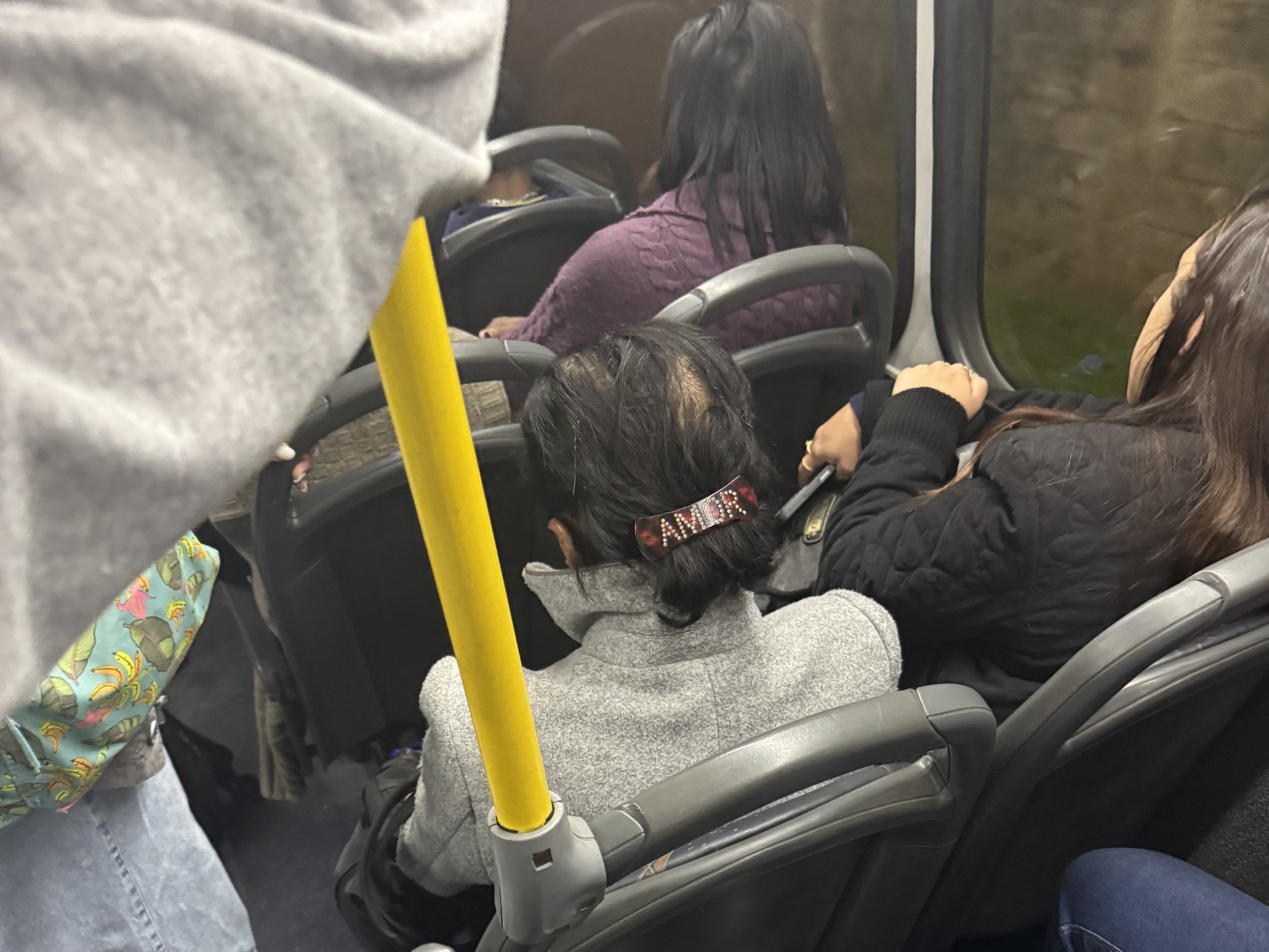 A woman with black hair sitting on a bus, wearing a purple sweater and looking out the window. Other passengers are visible, including a person with a colorful backpack and another with a hoodie, and a yellow vertical pole in the foreground.