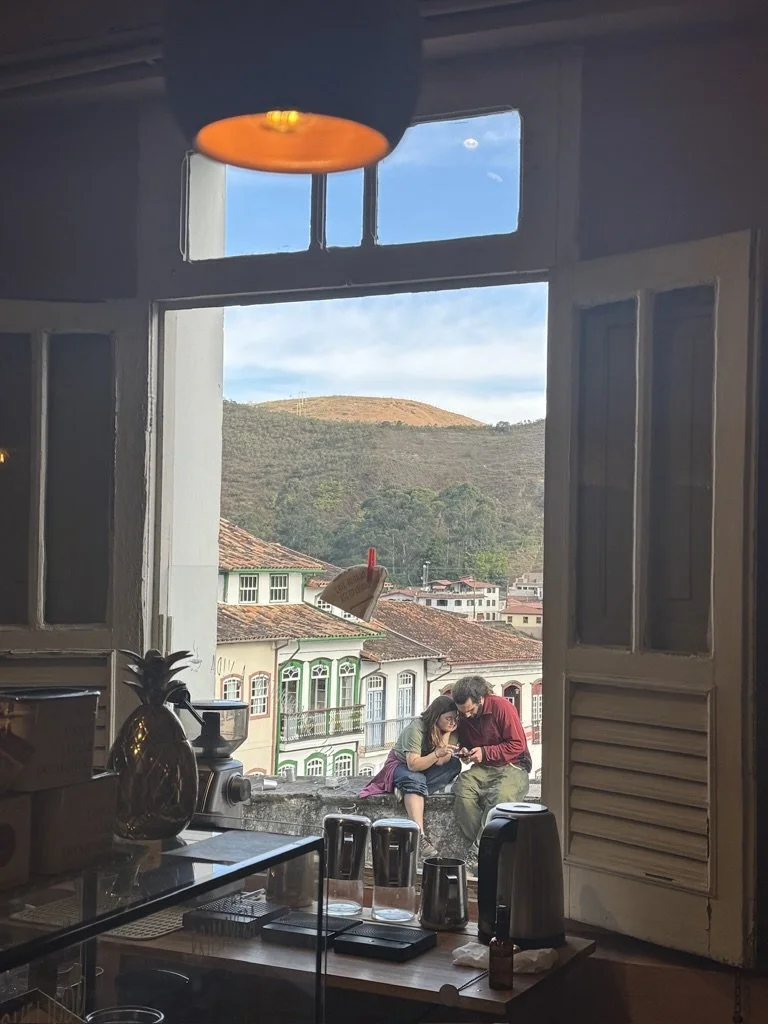 Two people sitting on a windowsill overlooking a neighborhood with colorful houses and a hill in the background, inside a kitchen with a pineapple-shaped container and kitchen appliances on the counter.