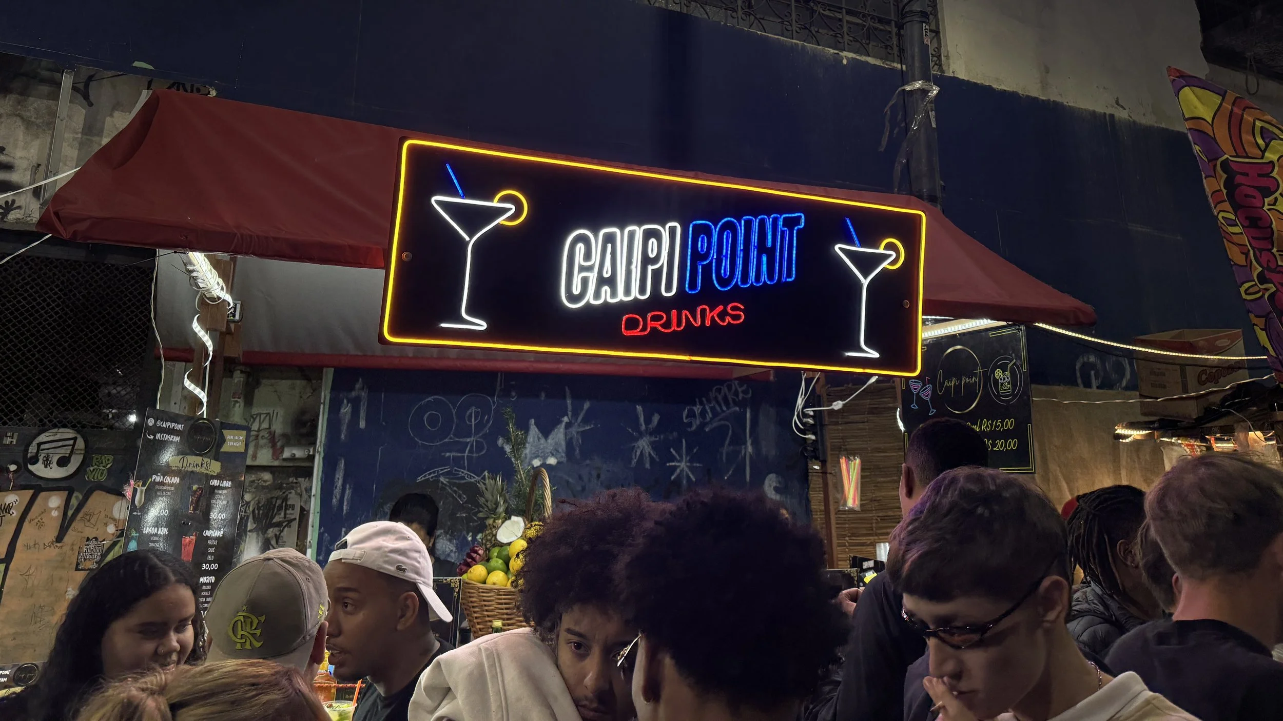 Neon sign for 'Cargo Point Drinks' with illustrations of martini glasses, set against a busy crowd scene at night under a canopy with graffiti and colorful decorations.