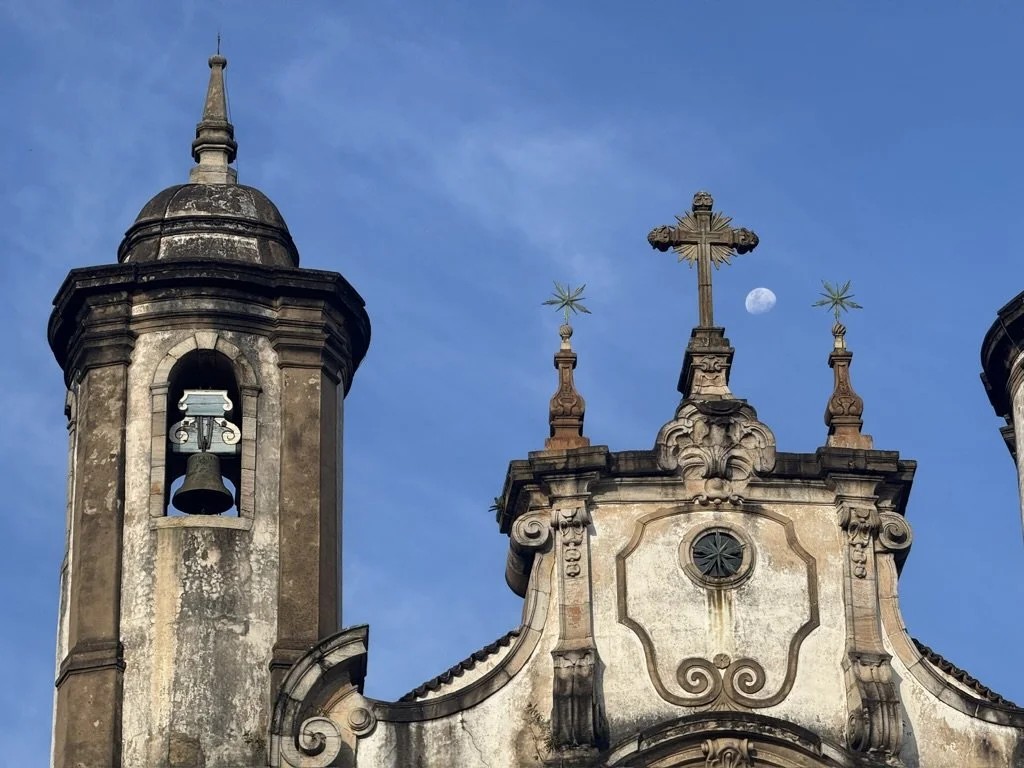 Close-up of a historic church with weathered stone architecture, topped with a cross, against a blue sky with the moon visible.