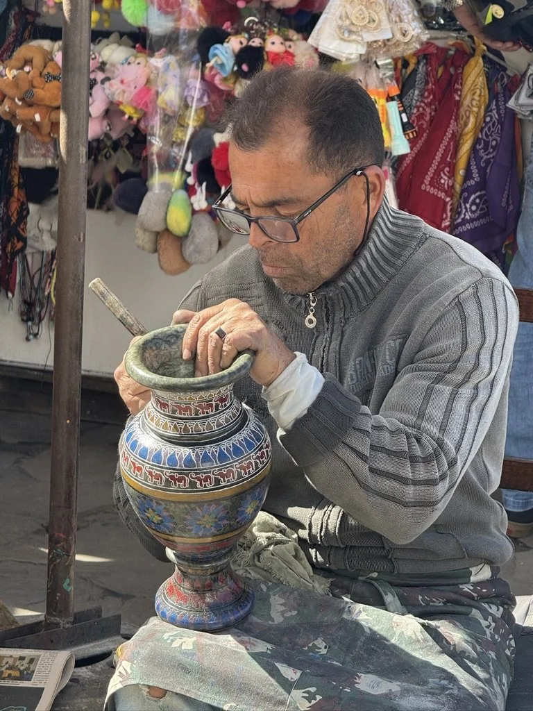 A man with glasses working on a colorful decorative vase at an outdoor market, with plush toys and fabrics in the background.