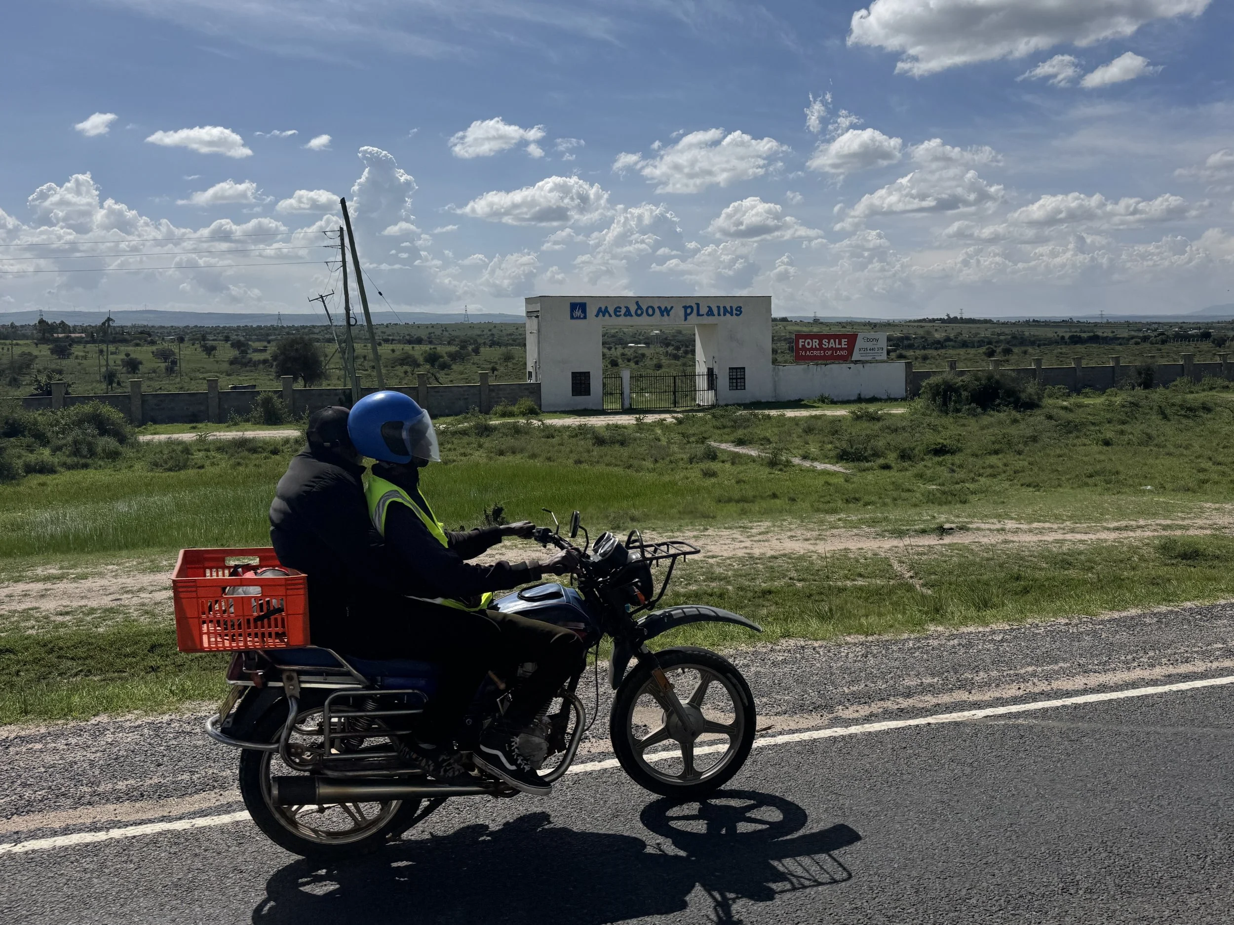 A person riding a motorcycle on a road with a red basket on the back, wearing a helmet and a reflective vest, with a landscape of green fields and a sign that reads 'Meadow Plains' and 'For Sale 74 Acres of Land' in the background, under a partly clo