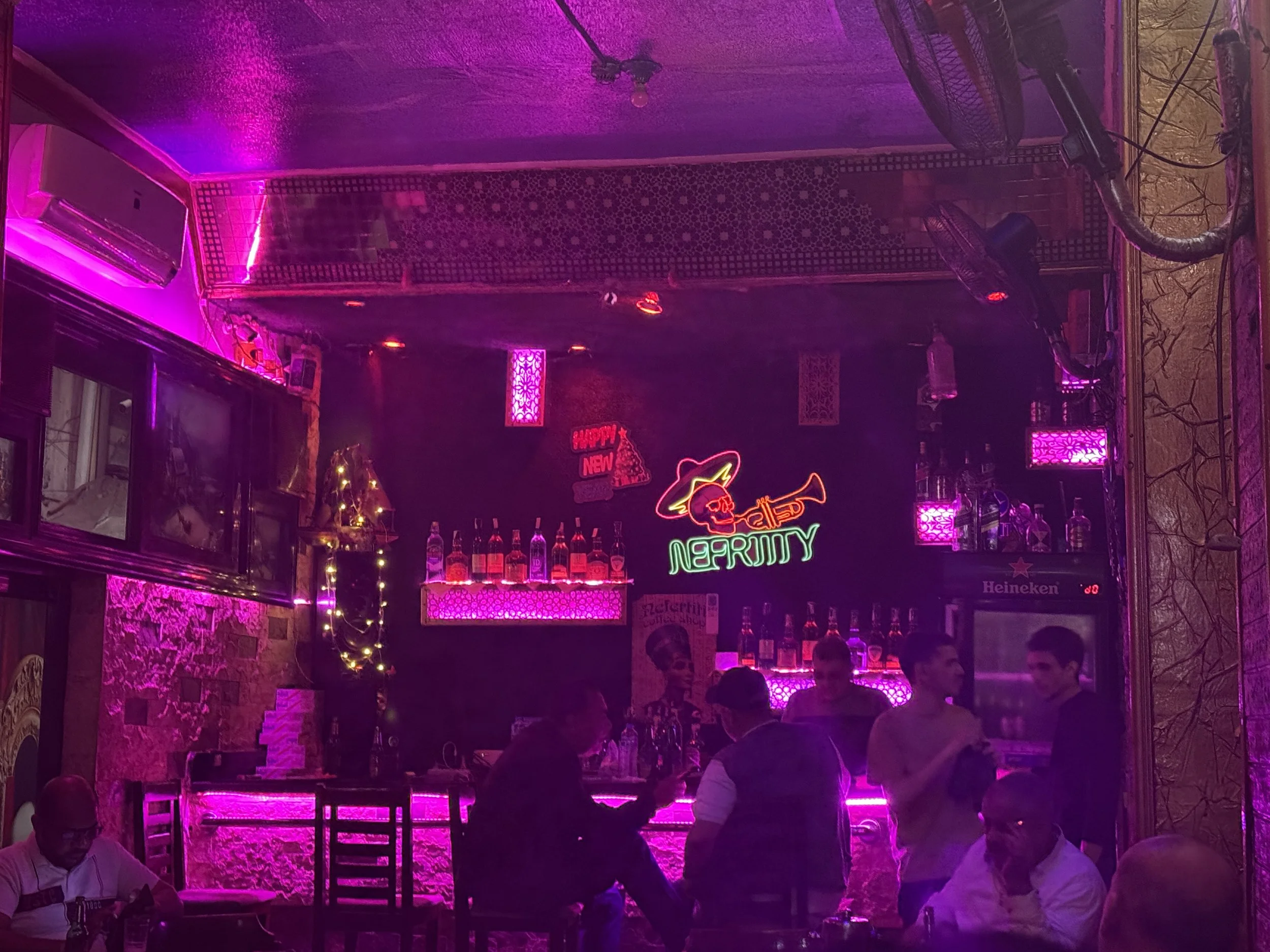 A bar or club interior illuminated with pink neon lights, with bottles of alcohol on display behind the bar, and people sitting and standing around enjoying drinks and conversation.
