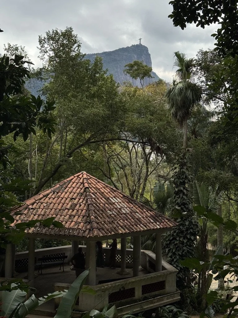 A park with a small round gazebo with a red-tiled roof surrounded by trees and foliage, with the Christ the Redeemer statue atop Corcovado Mountain in the background.