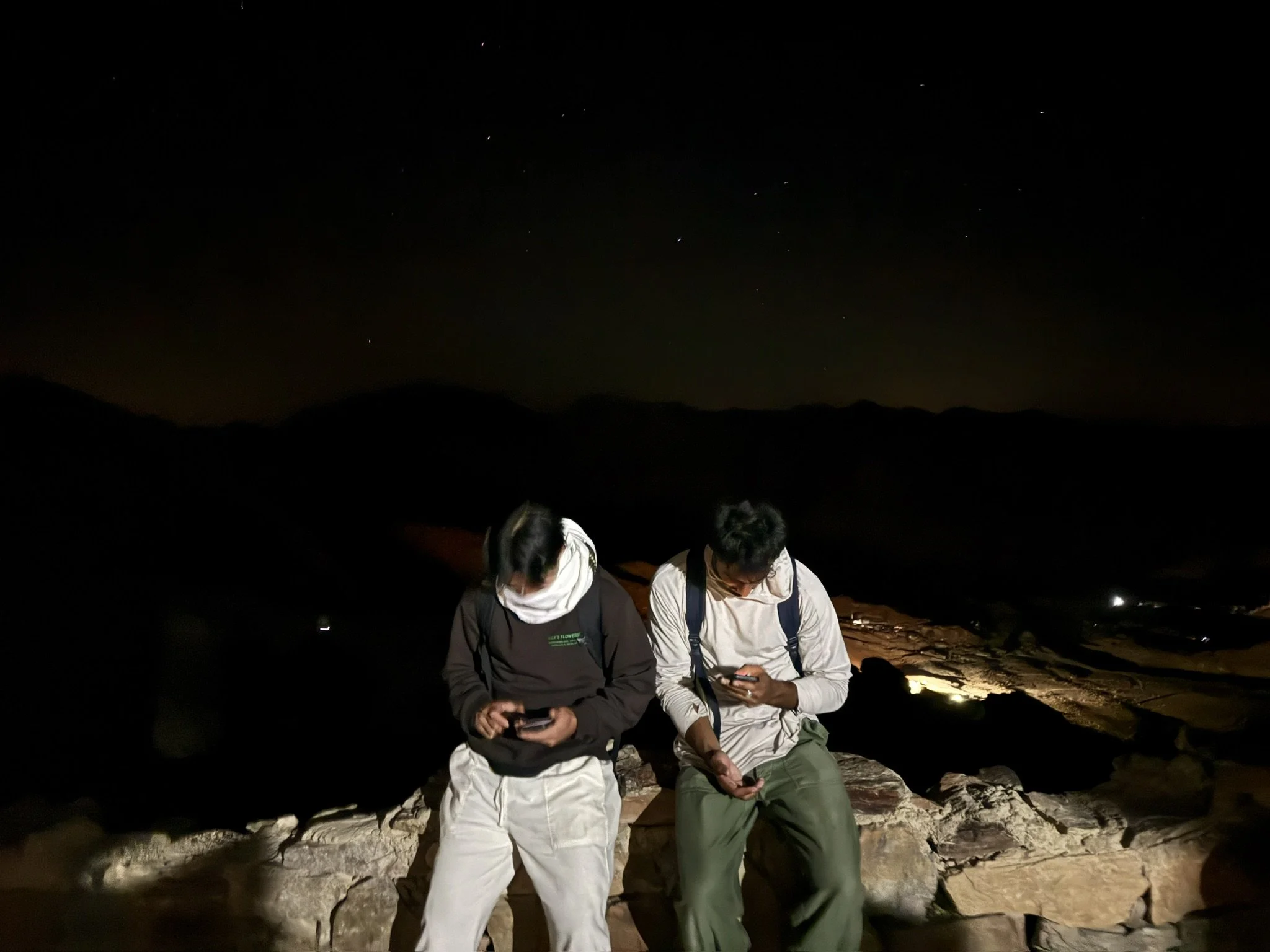 Two people sitting on rocks at night, looking at their phones with a dark sky and stars above.