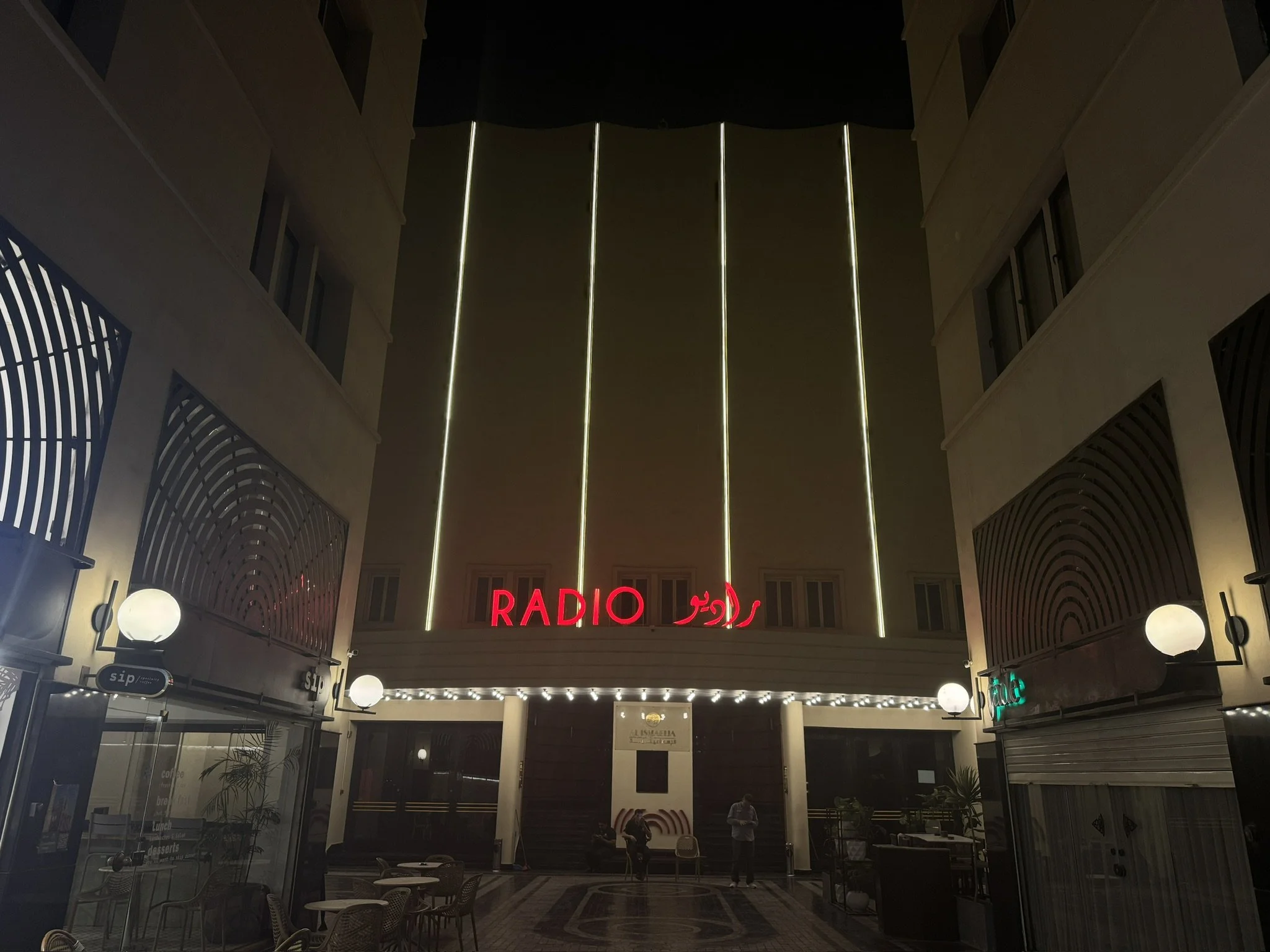 Night view of a shopping mall or hotel atrium with a neon sign that reads 'RADIO' in red. The atrium features modern design elements, with curved metal railings, circular wall lamps, and a high ceiling with vertical light strips. There are a few peop