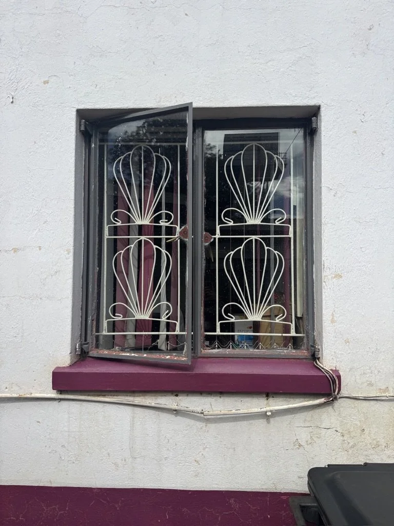 A window with a decorative iron grille on an exterior wall, one pane open, with purple window sill and trim, and a black object partially visible at bottom right.