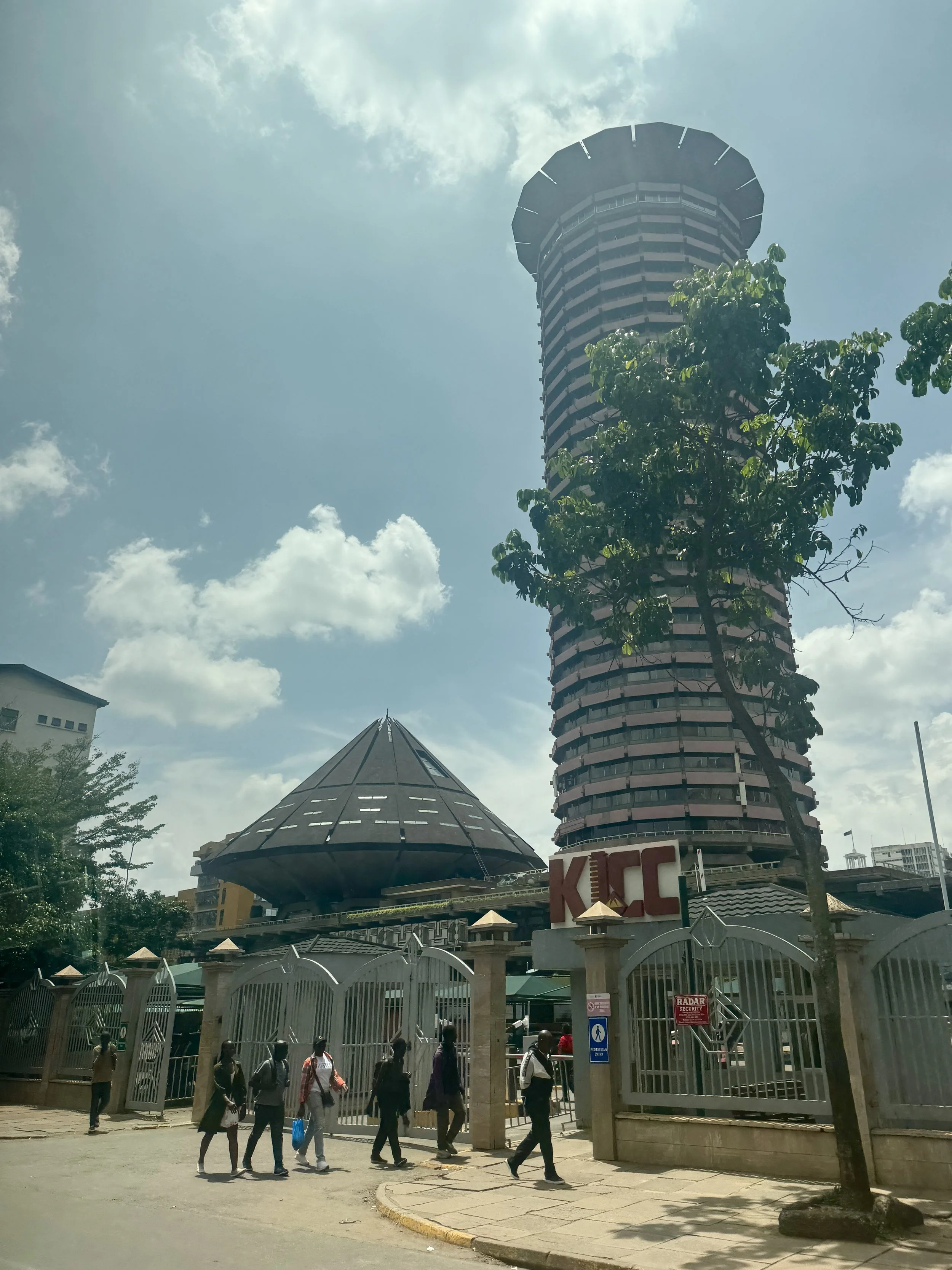 A group of people walking past a gated entrance with a KFC sign, with a tall, uniquely designed tower building in the background and a smaller, circular structure with a roof resembling a traditional hat.