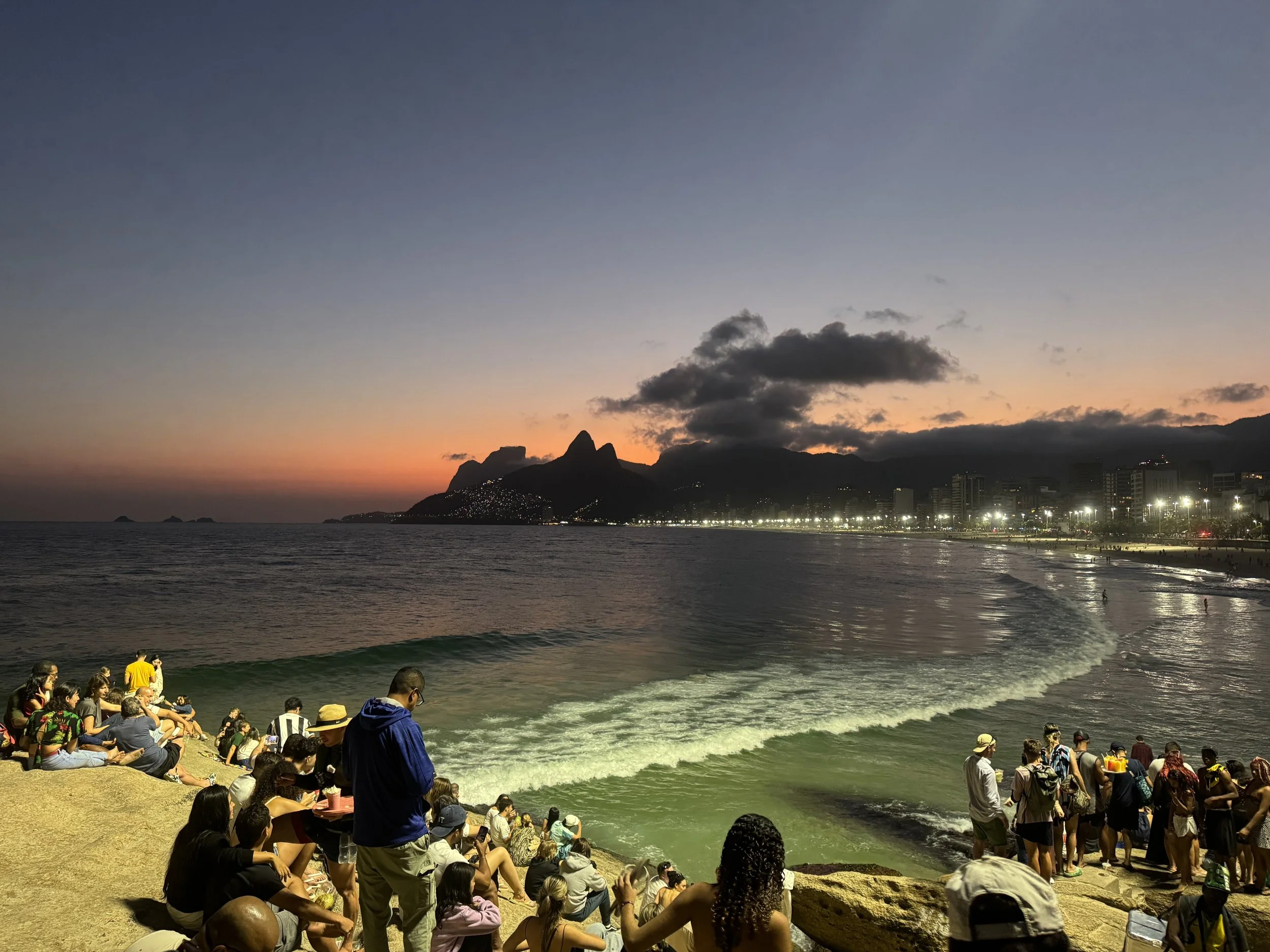 People sitting and standing on a sandy beach at sunset, looking at the ocean waves, with a cityscape and mountains in the background.