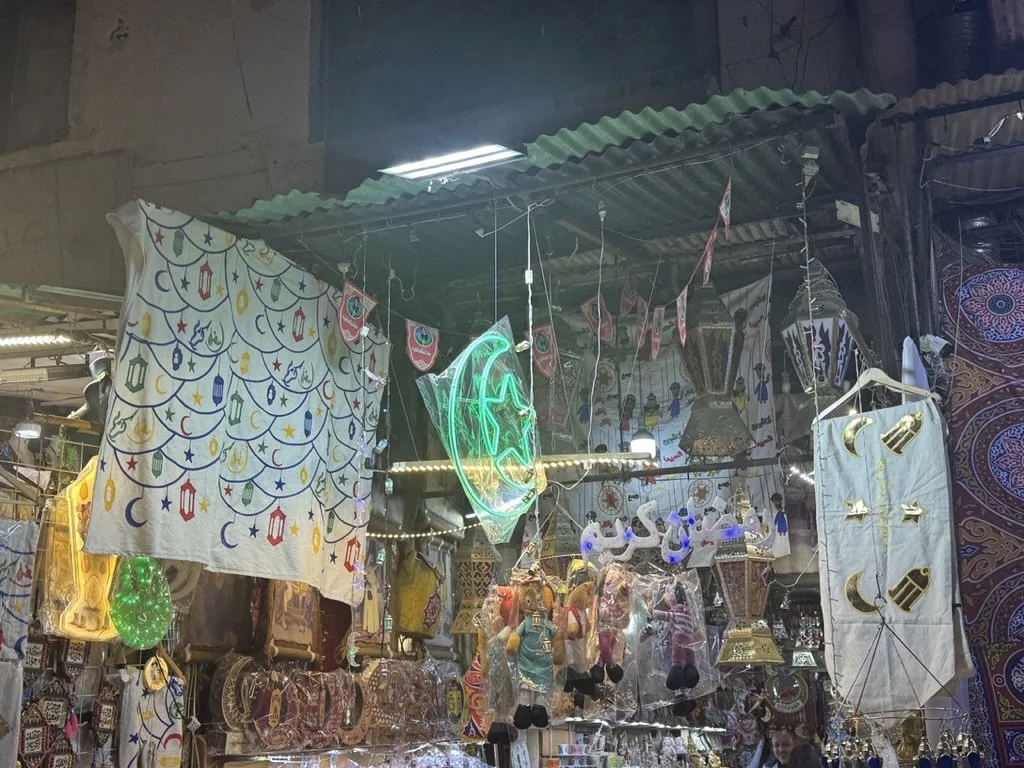 A market stall adorned with Islamic-themed decorations, including a neon green crescent moon and star sign, hanging lanterns, and Islamic symbols on fabrics. The stall displays various items like hanging ornaments and textiles.