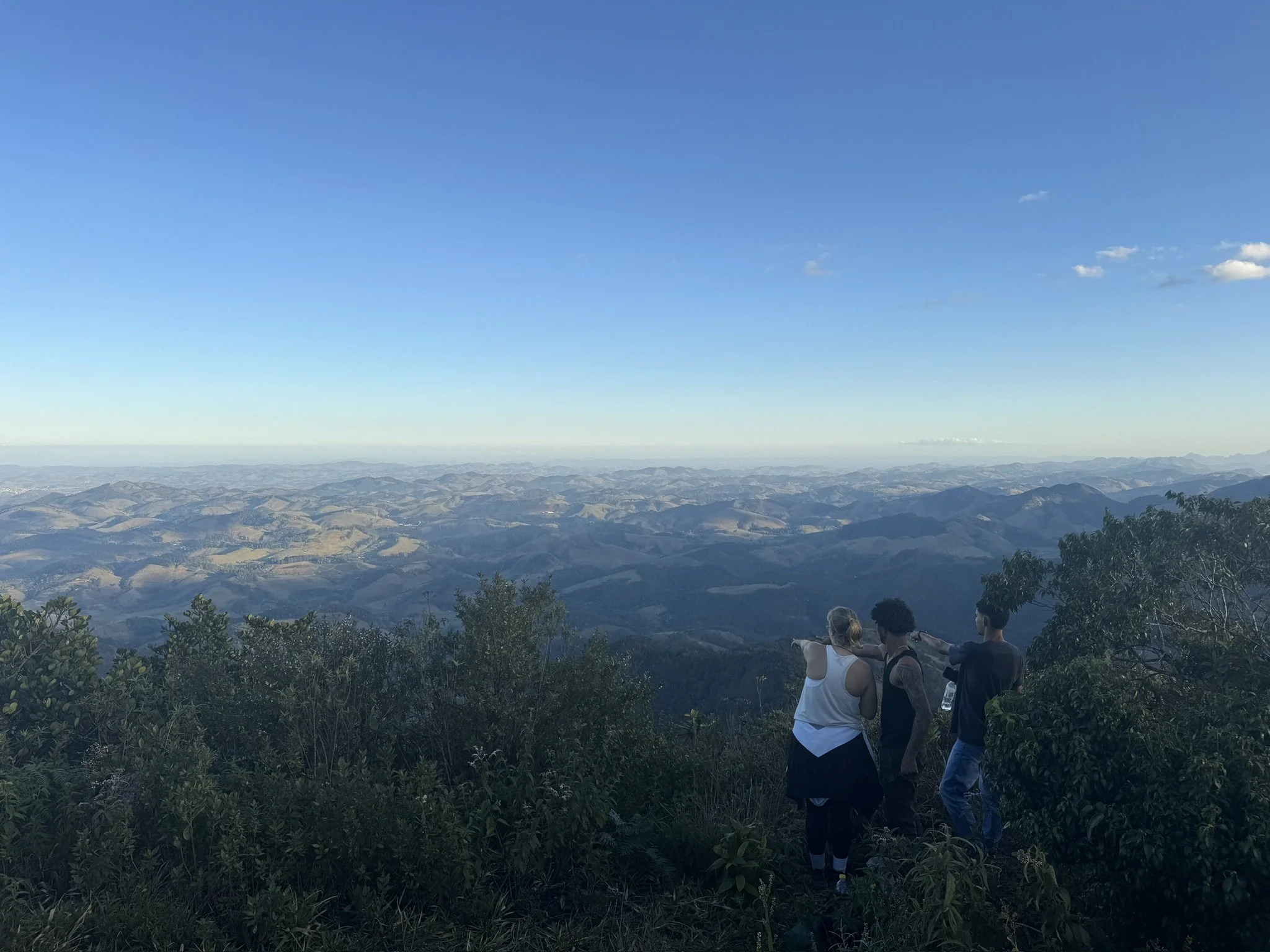 Three people standing on a mountain overlook, gazing at a vast landscape of rolling hills and mountains under a clear blue sky.