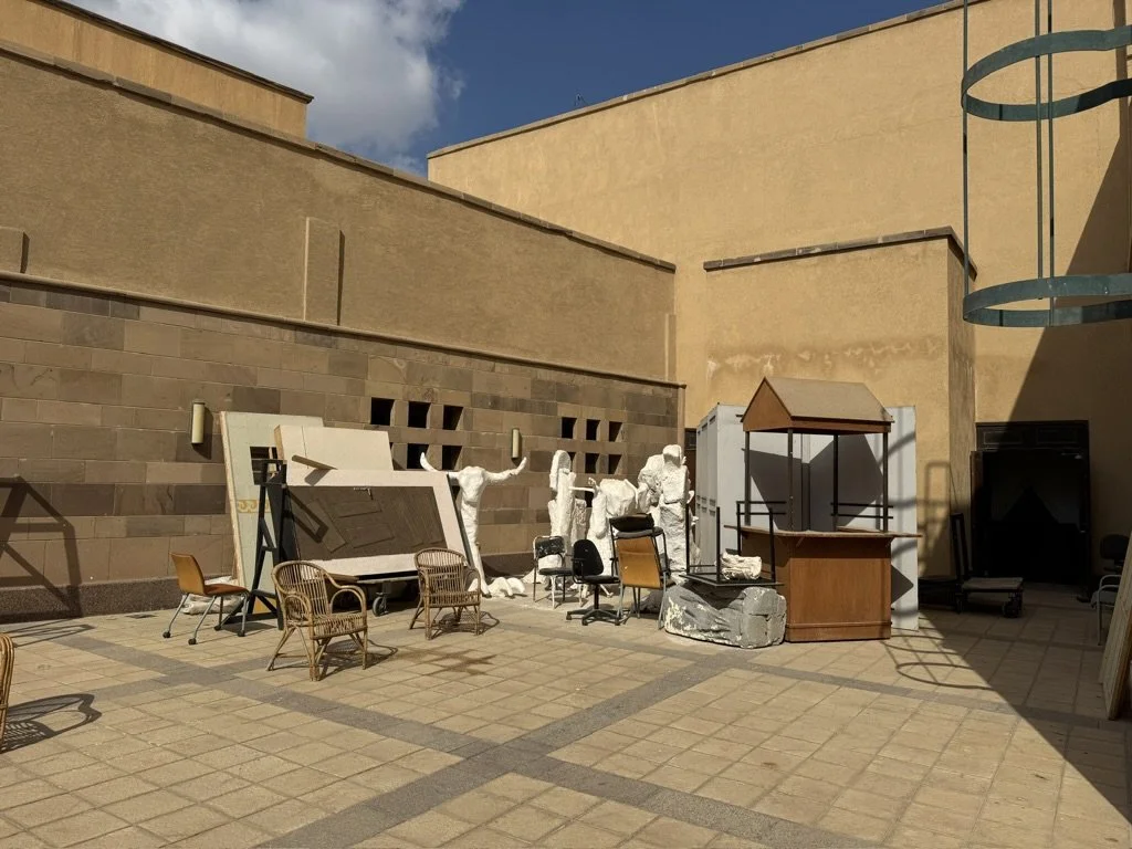 Outdoor courtyard scene with various furniture and sculptures, including chairs, tables, and white statues of people, against a beige building with a blue sky background.