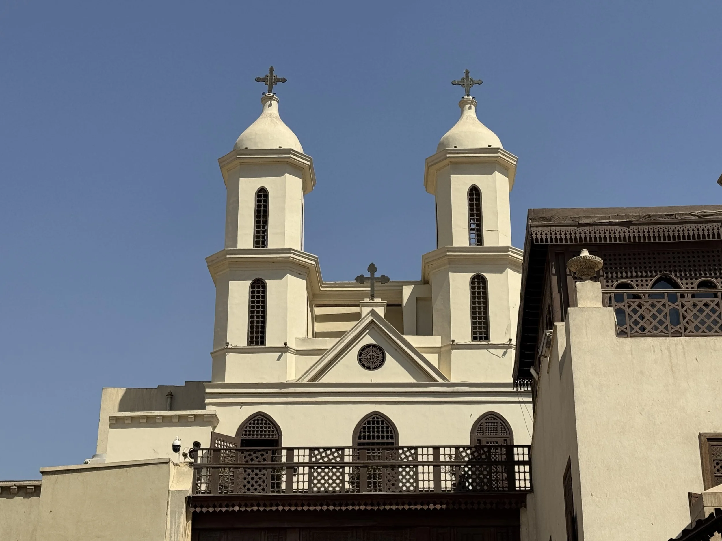 The image shows the upper section of a white church with two tall bell towers topped with crosses, set against a clear blue sky. Part of the building's facade and a balcony with decorative railing are visible.