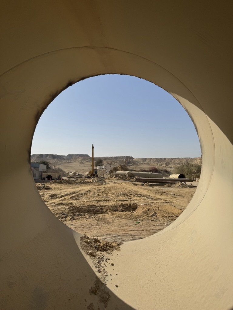 View of a construction site through a large pipe with a circular opening, showing dirt roads, cranes, and industrial equipment in a desert landscape under a clear sky.