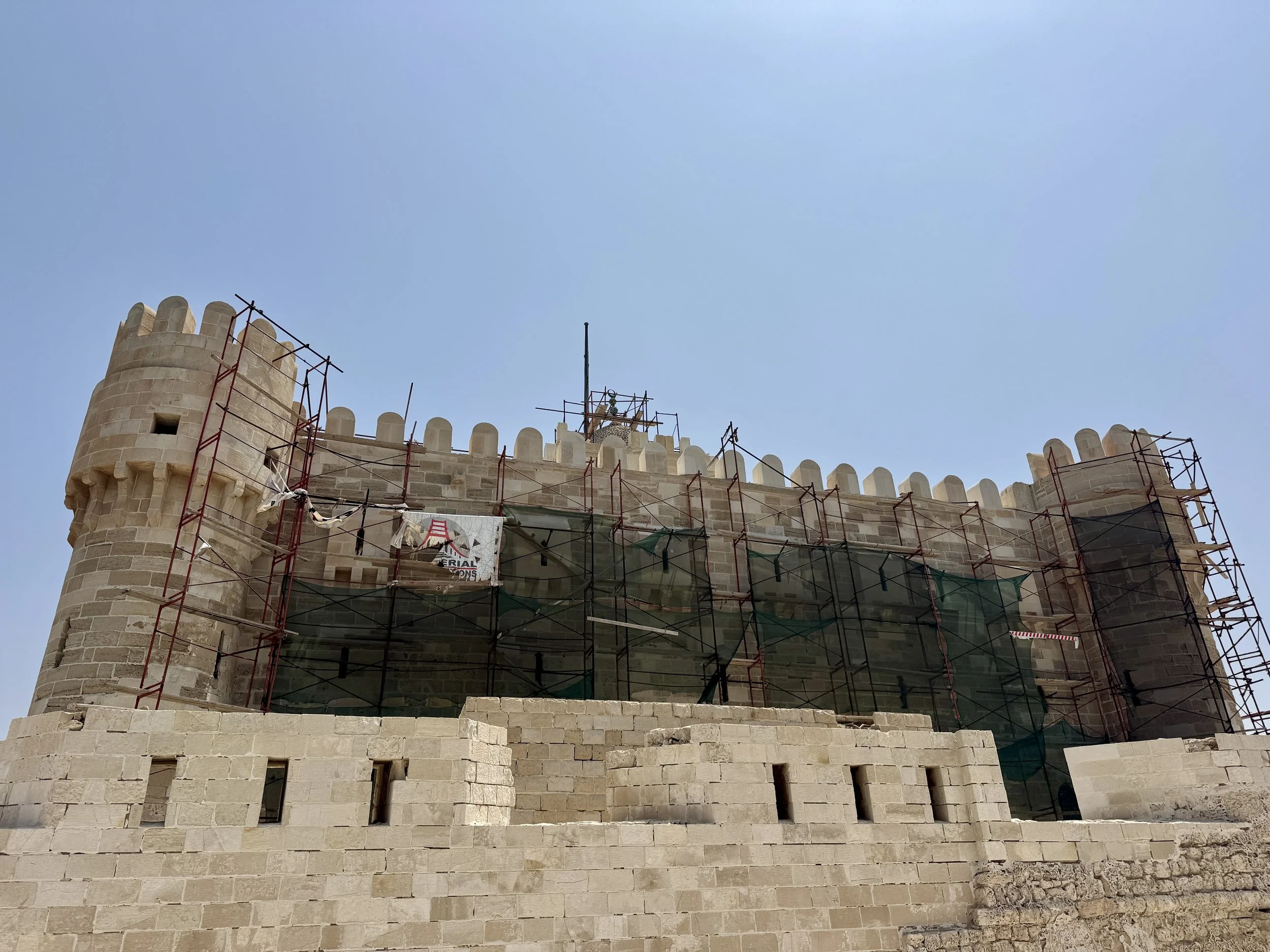 The image shows a historical stone castle under renovation with scaffolding on the walls and towers, set against a clear blue sky.