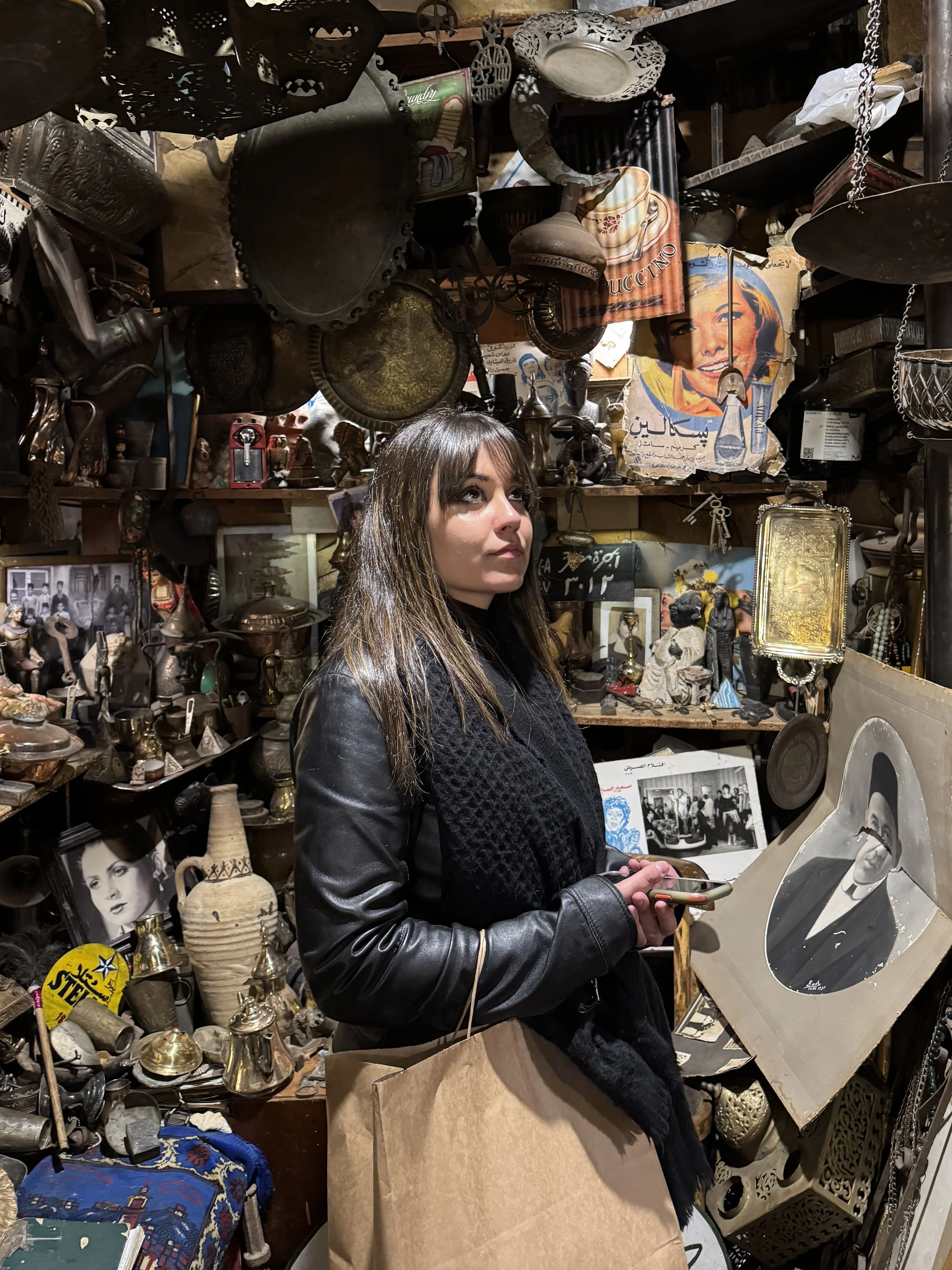 A woman with long brown hair, wearing a black leather jacket, is in an antique shop looking at various vintage items. She is holding a phone and a shopping bag, surrounded by old photographs, sculptures, and metalware, with shelves filled with collec