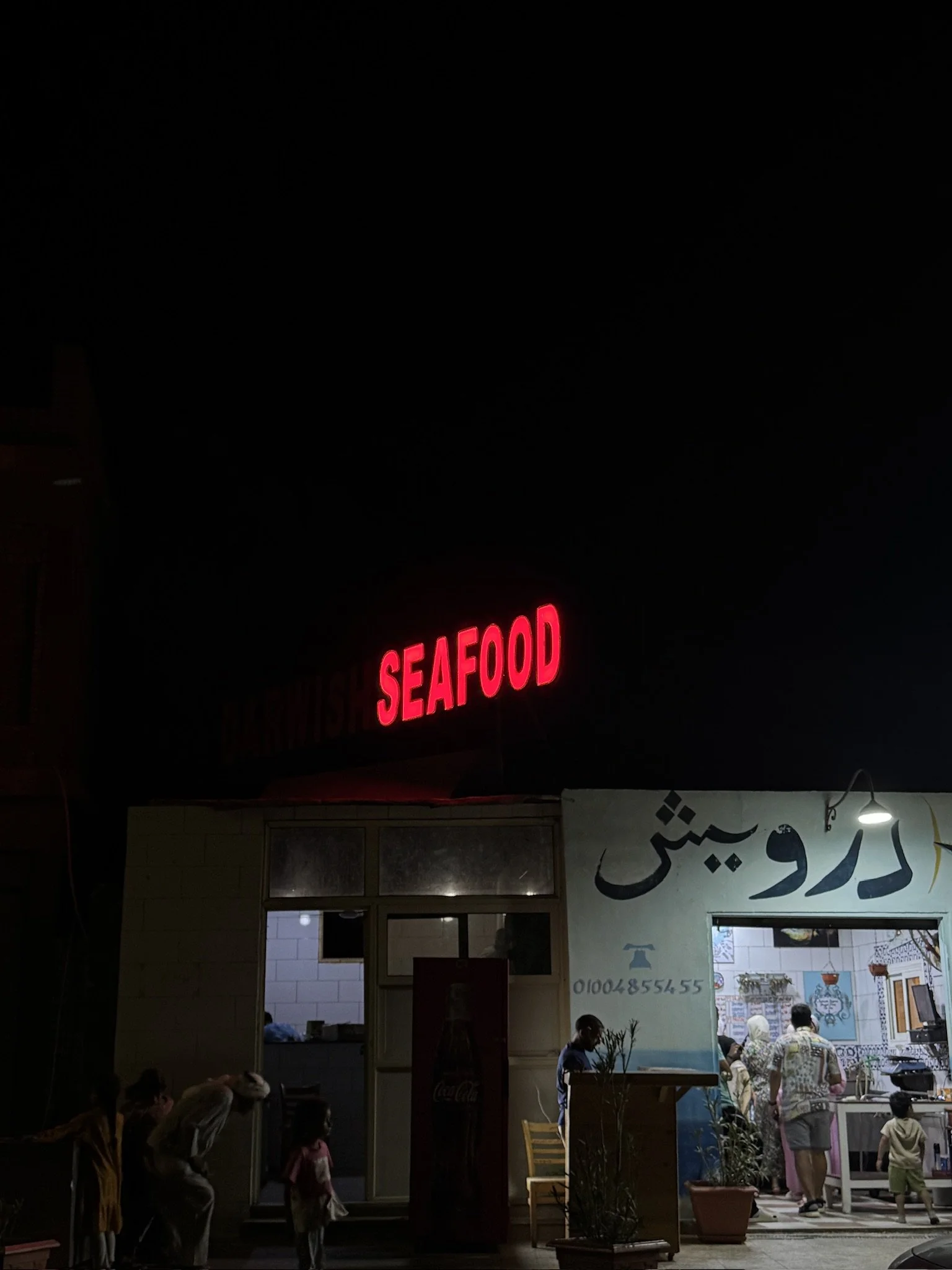 People standing outside a seafood restaurant at night, with a bright red neon sign reading 'SEAFOOD' and a large sign with Arabic text.