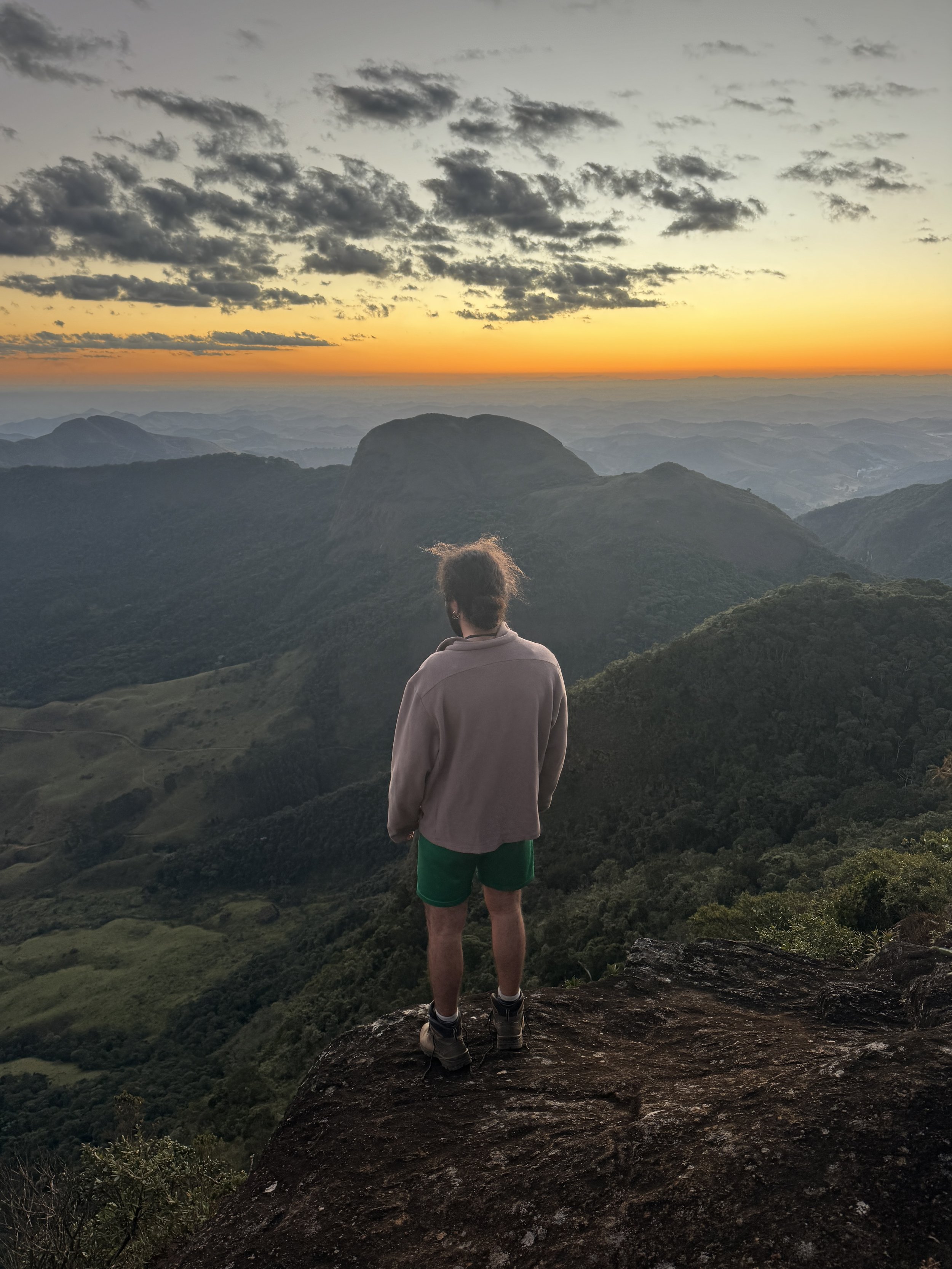 A person with curly hair wearing a hoodie and green shorts stands on a rocky ledge, overlooking a mountain landscape at sunset with a sky filled with clouds.