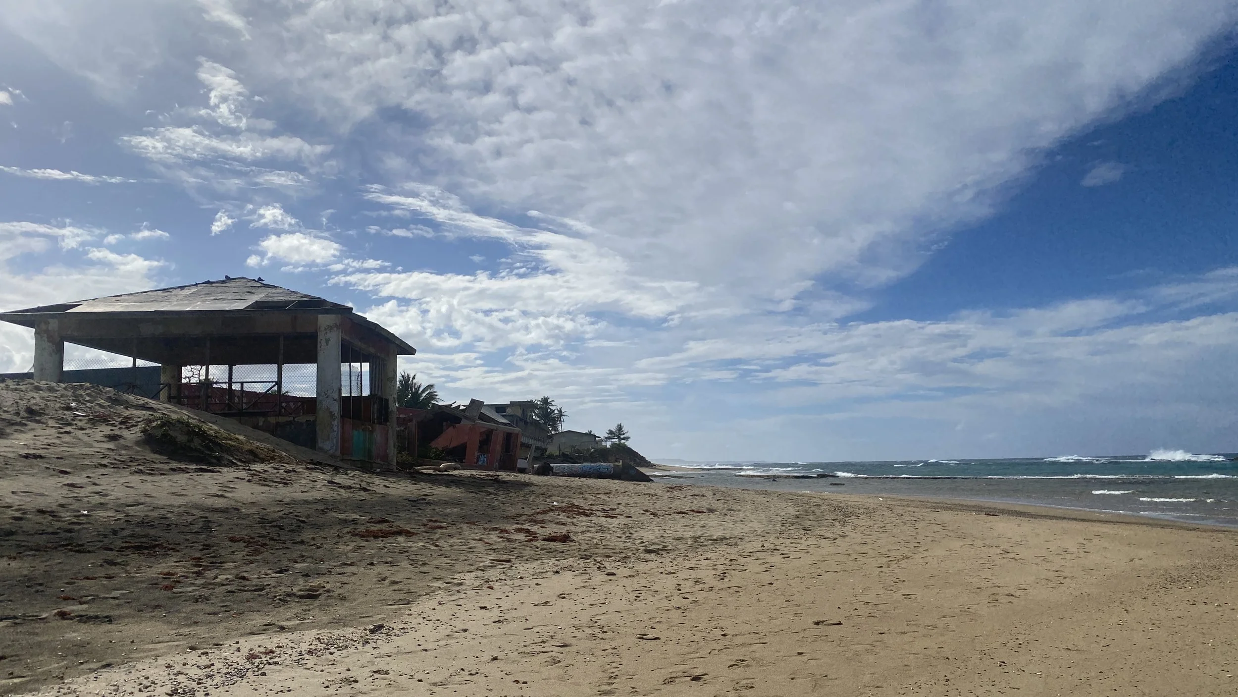 A beach scene featuring an abandoned, dilapidated building on the sand, with the ocean waves and a blue sky with clouds in the background.