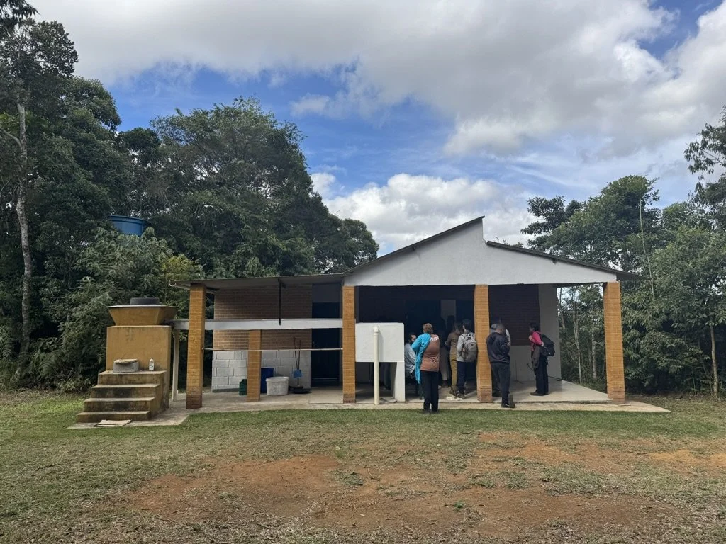 People standing outside a small building in a rural area, surrounded by trees and under a partly cloudy sky.